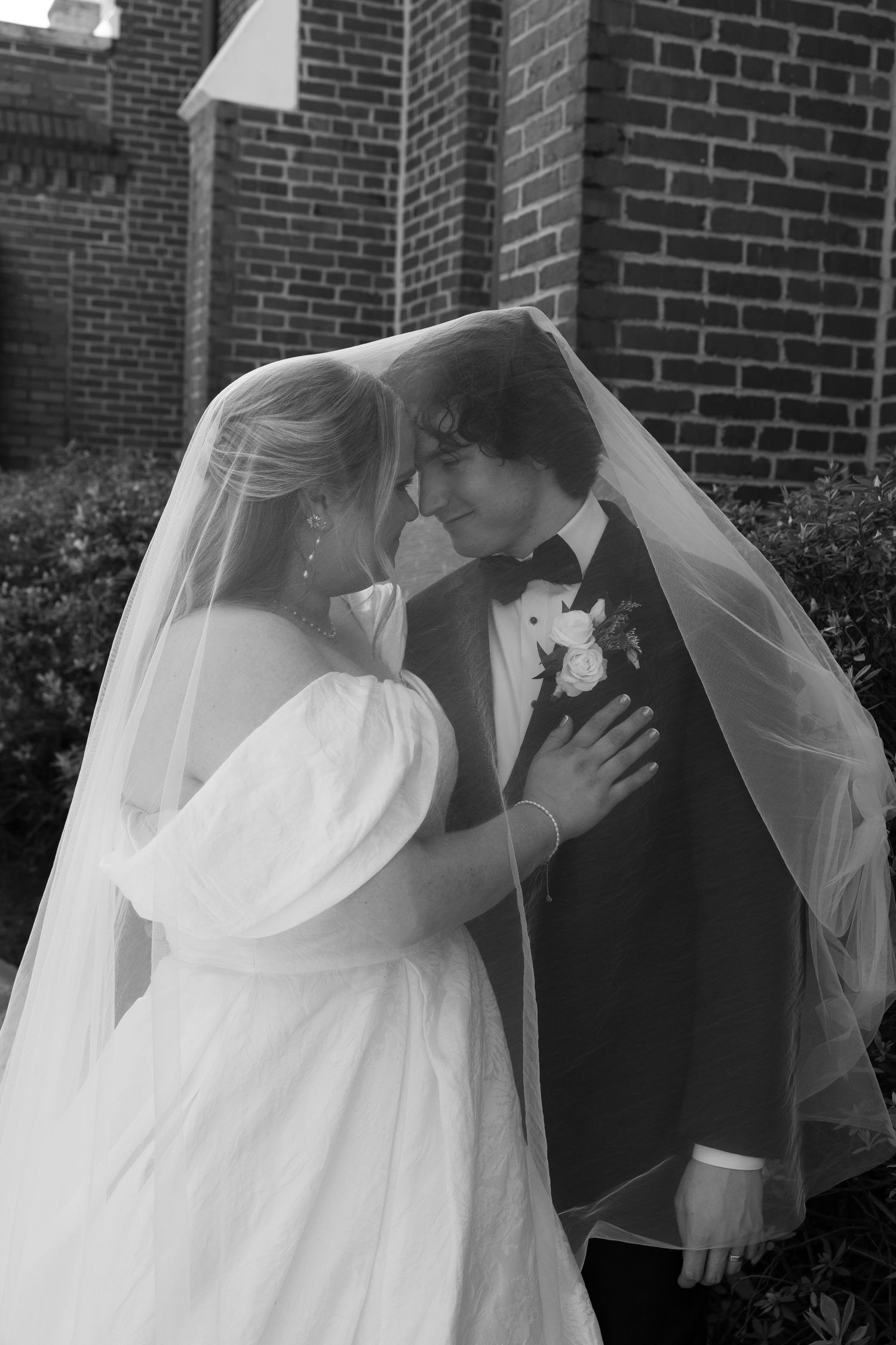 Black and white photo of bride and groom sharing romantic moment under wedding veil outside brick church.