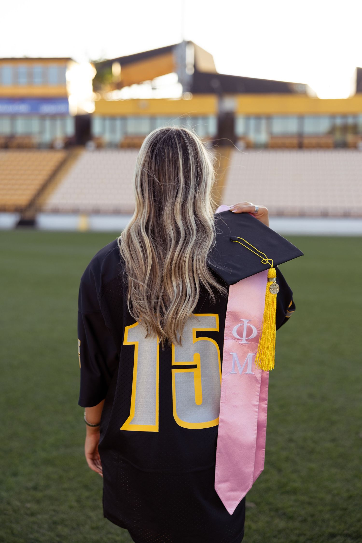 Back view of graduate in jersey number 15 holding cap with pink stole on football field.