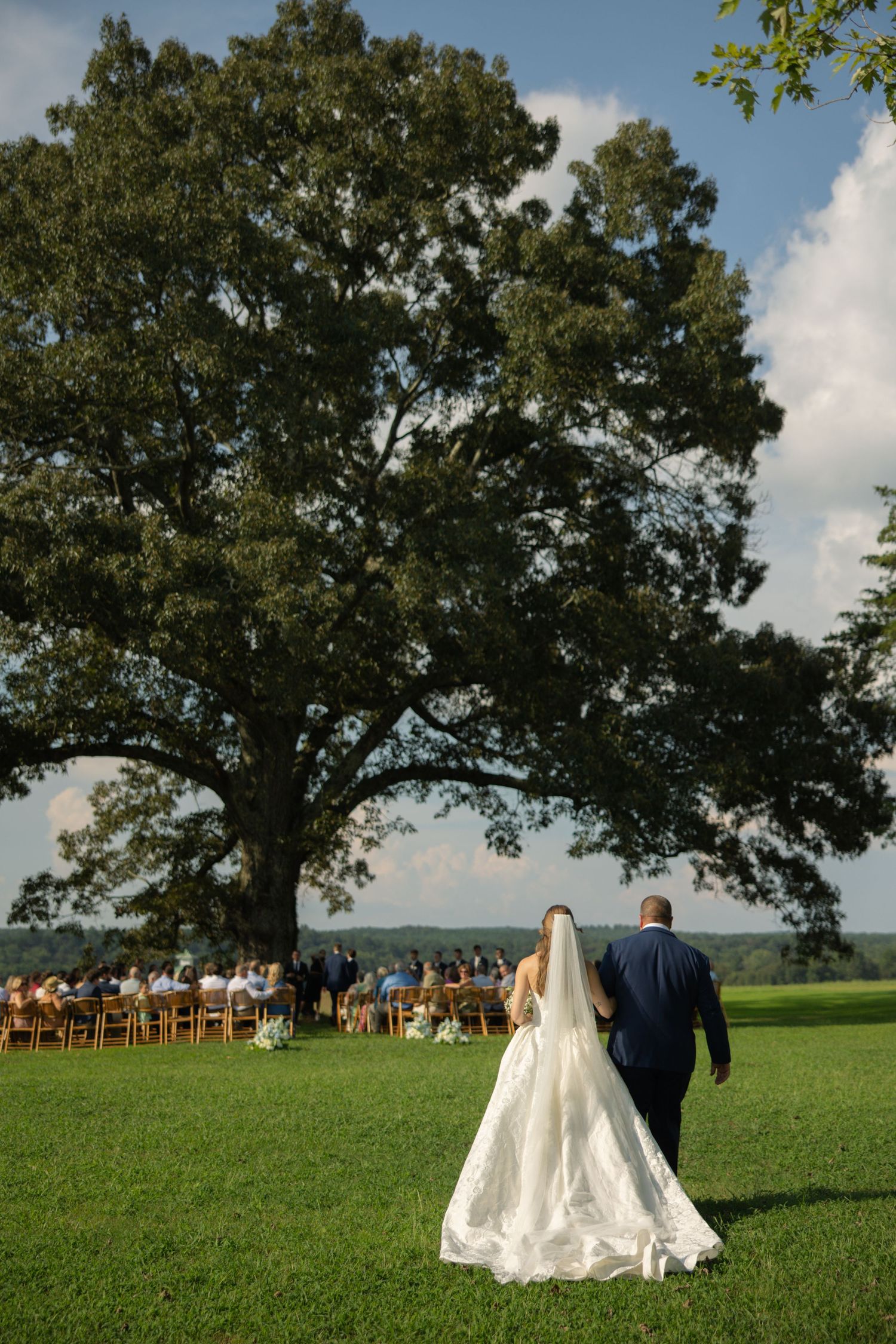 A couple walks across a lush green lawn with wedding guests seated in the background under blue skies.