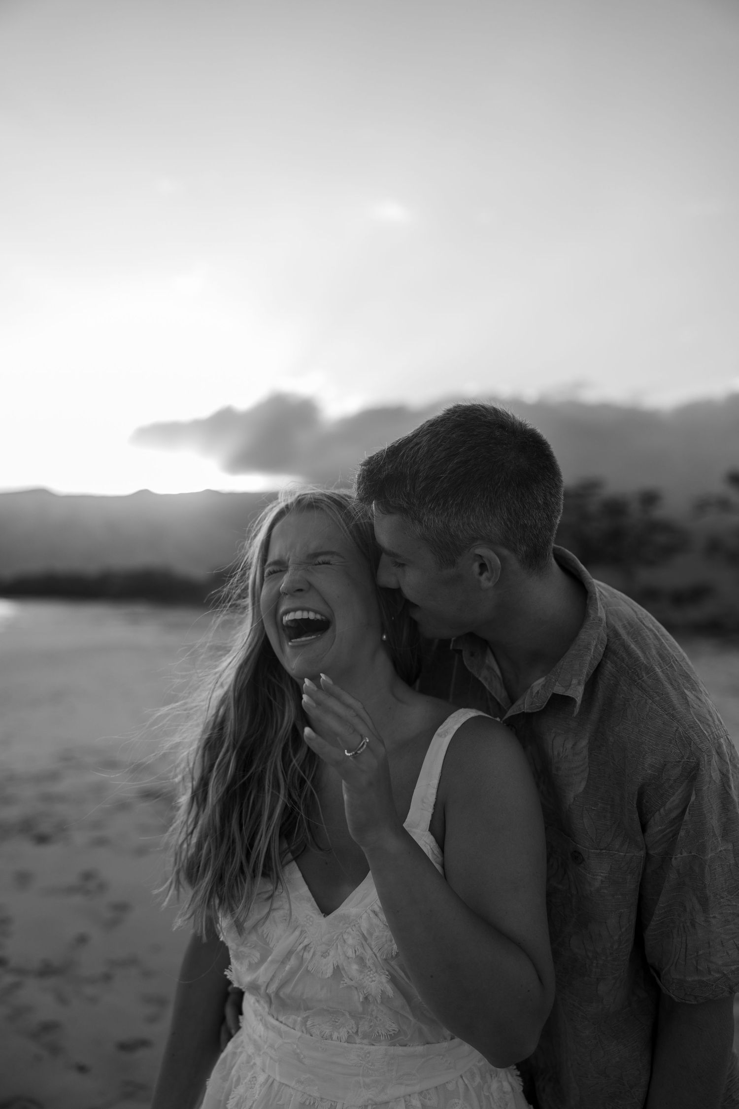 A candid black and white shot of two people sharing a joyful, intimate moment on the beach at sunset.