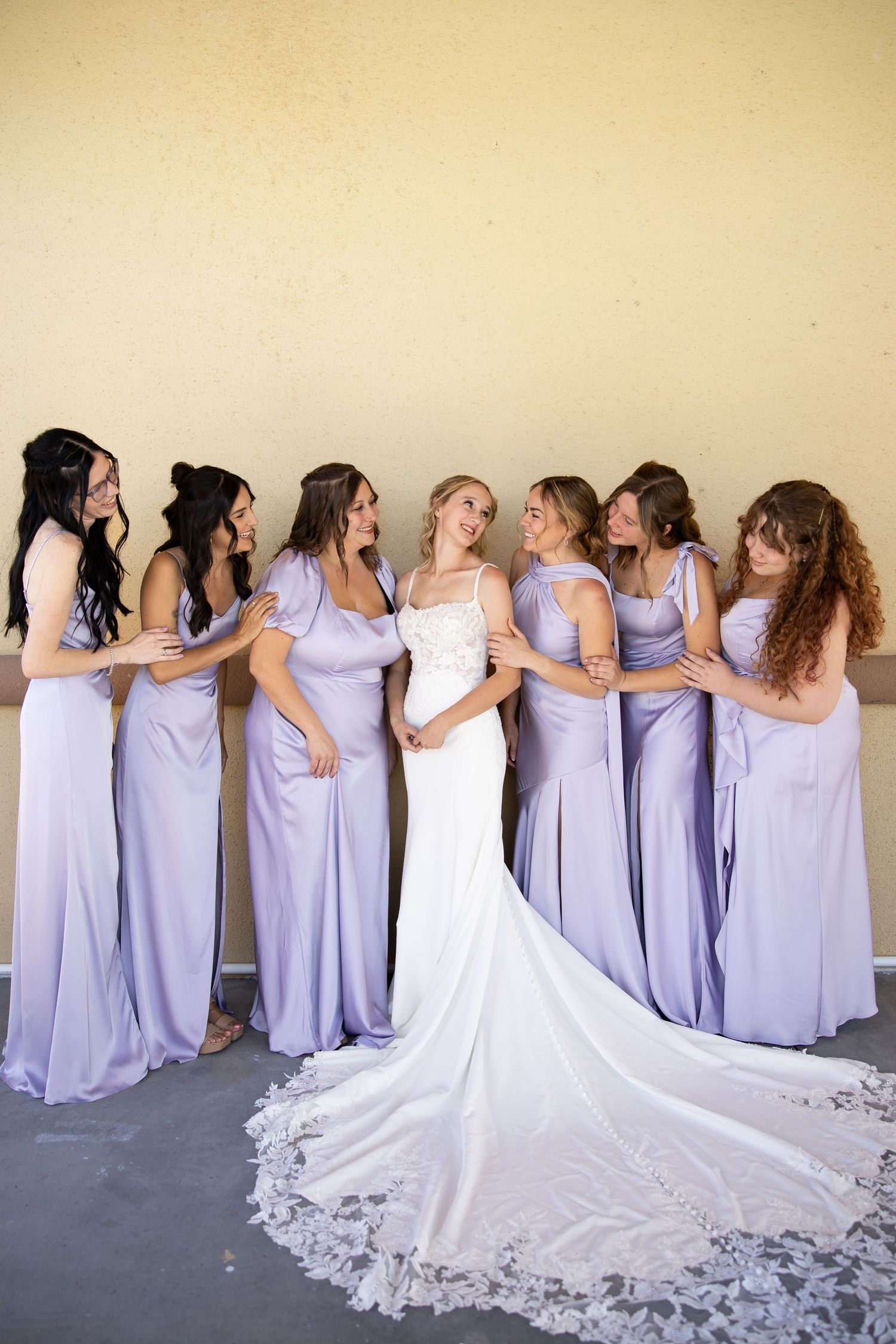 Bride in white gown surrounded by seven bridesmaids in lavender satin dresses, smiling together indoors.