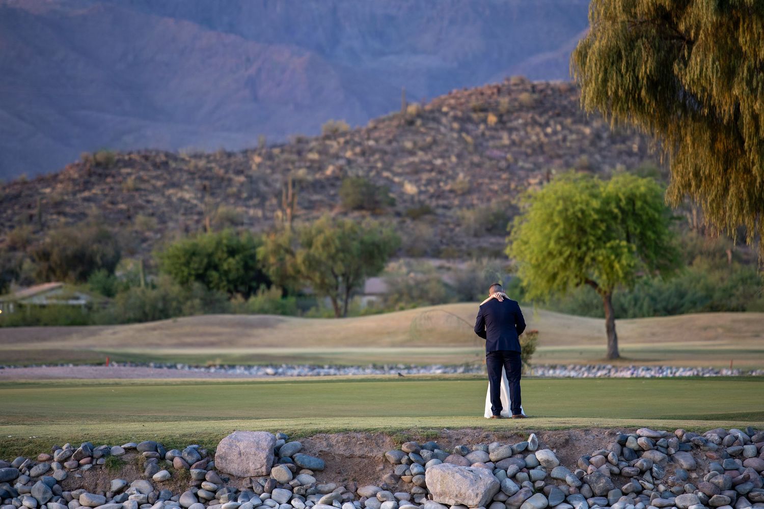 Golfer standing on lush green golf course with rocky landscape, trees, and mountain backdrop at sunset.