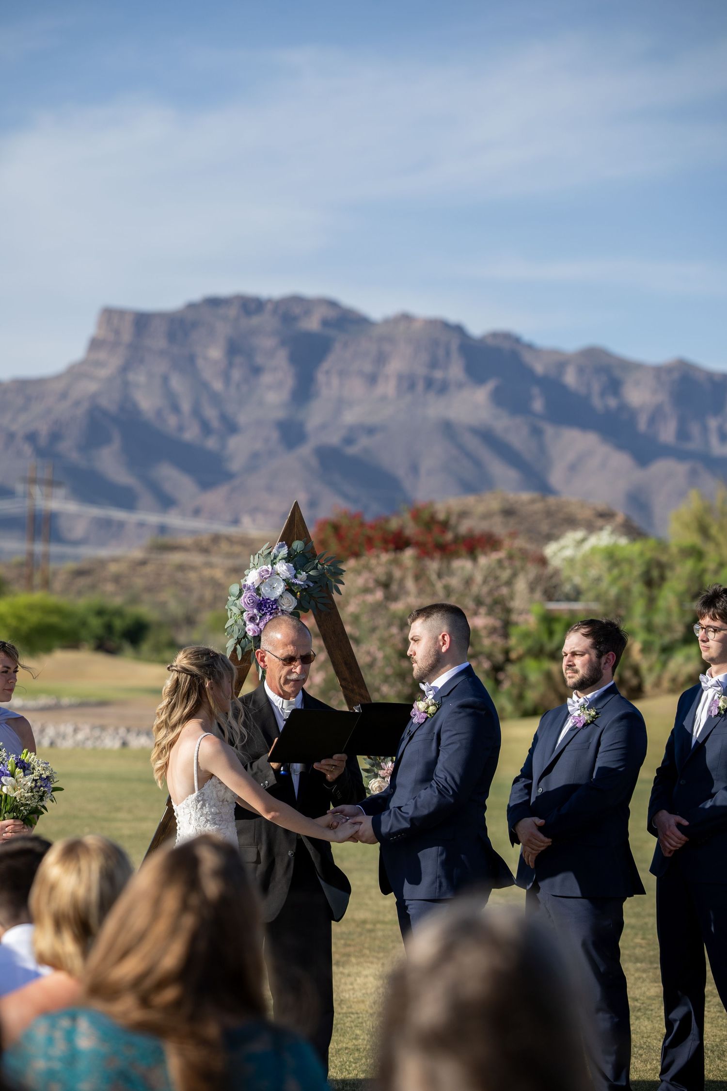 Outdoor wedding ceremony with couple exchanging vows before officiant, wooden arch with purple flowers, mountain backdrop.