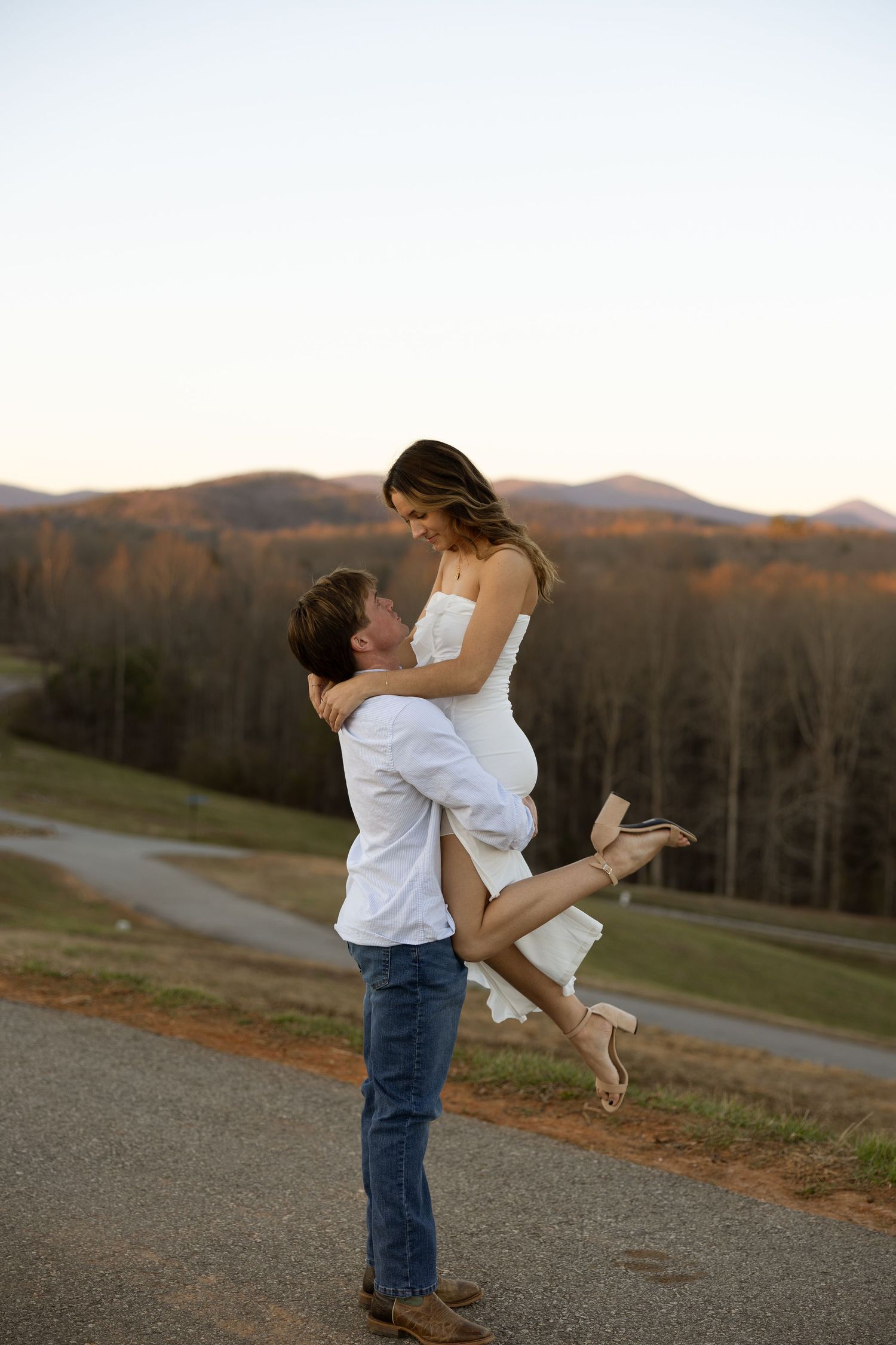 A playful couple dances together at sunset on a hilltop overlooking mountains and autumn trees.