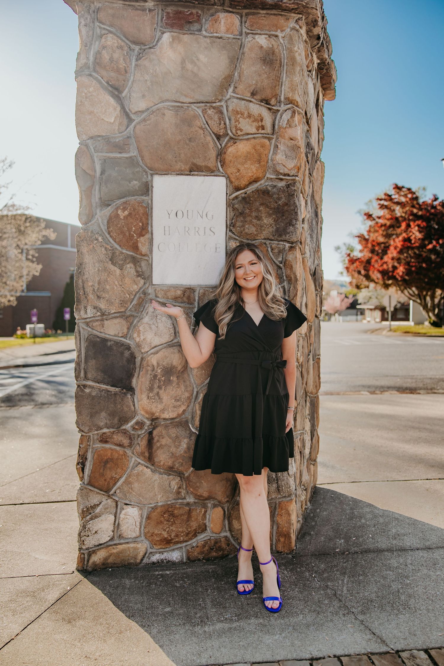 Graduate in black dress poses against stone wall with purple heels.