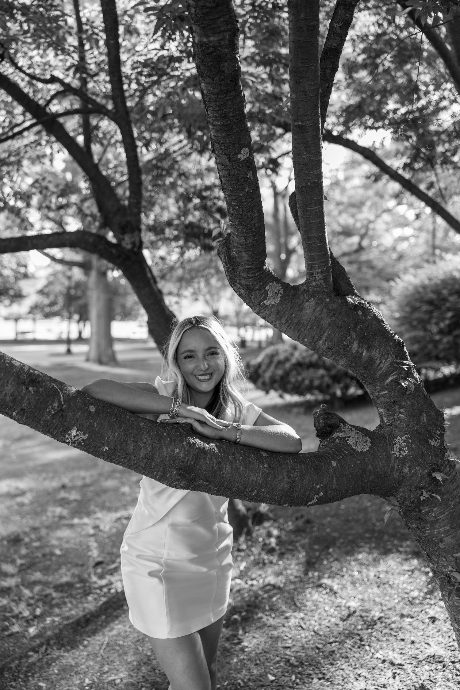 Black and white photo of someone posing casually by a large tree trunk on campus.