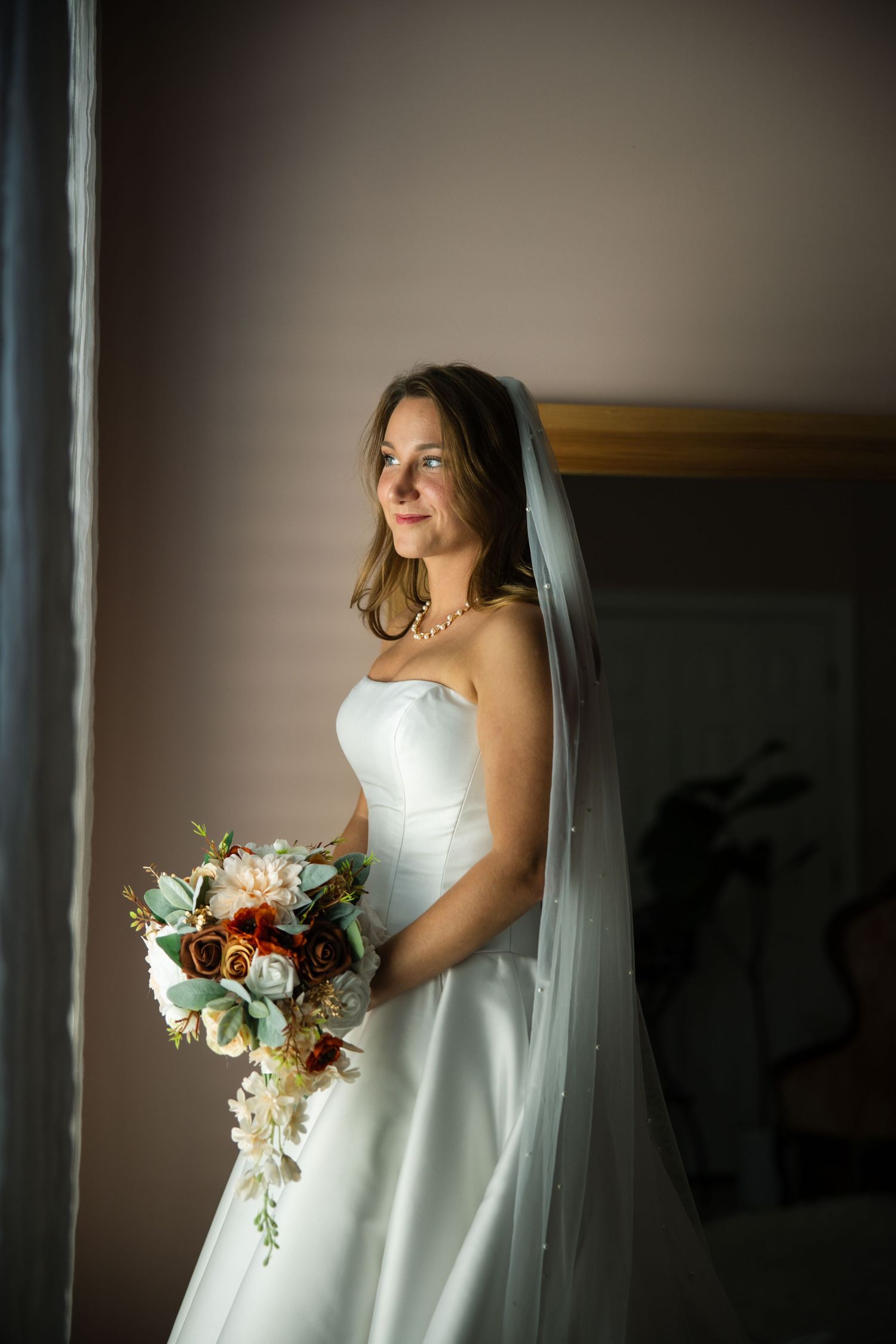 A bride in a white wedding gown stands in dramatic window light holding a bouquet of mixed flowers.