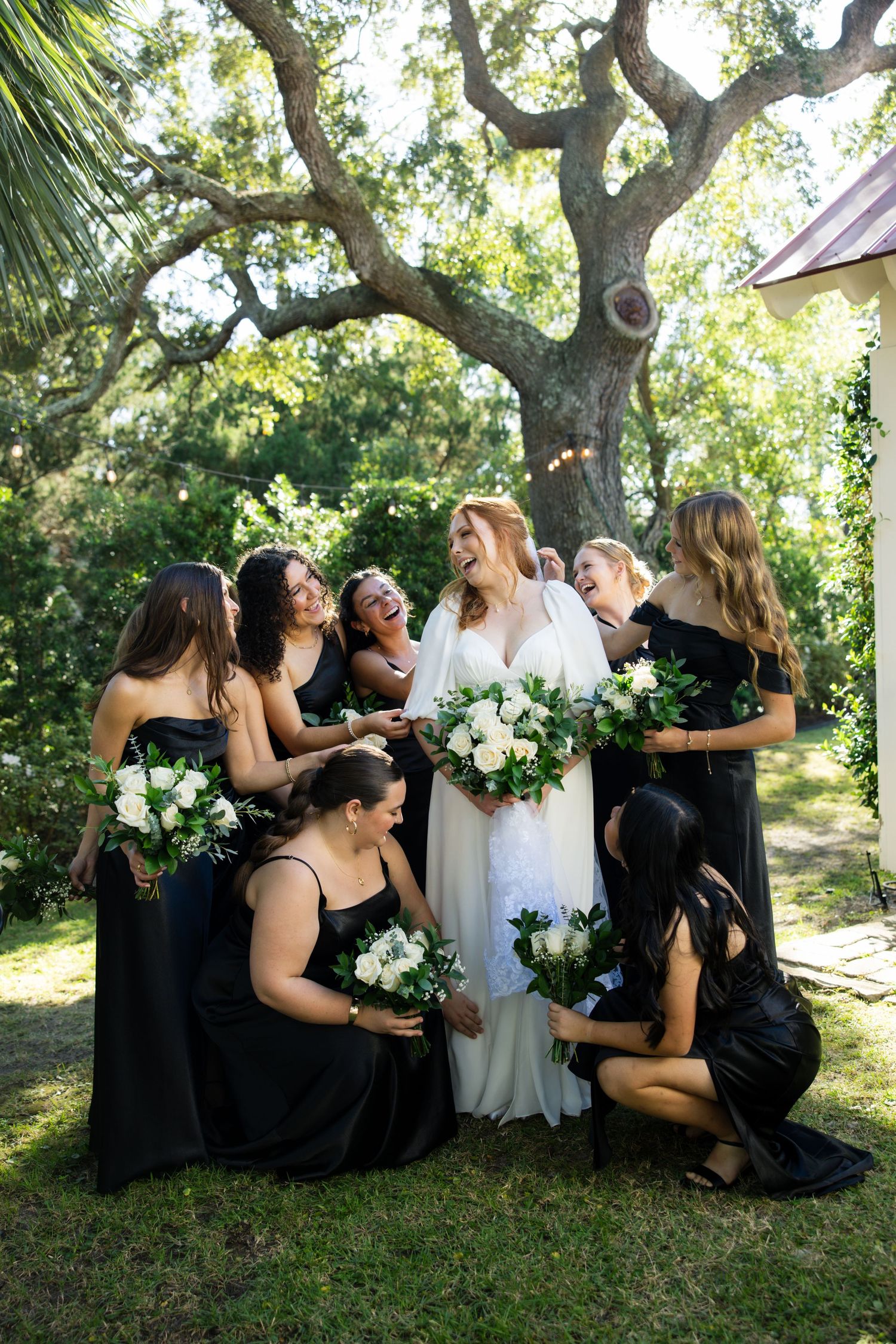 A bridal party in black dresses and white gown sits together on grass under a shady tree during an outdoor wedding.