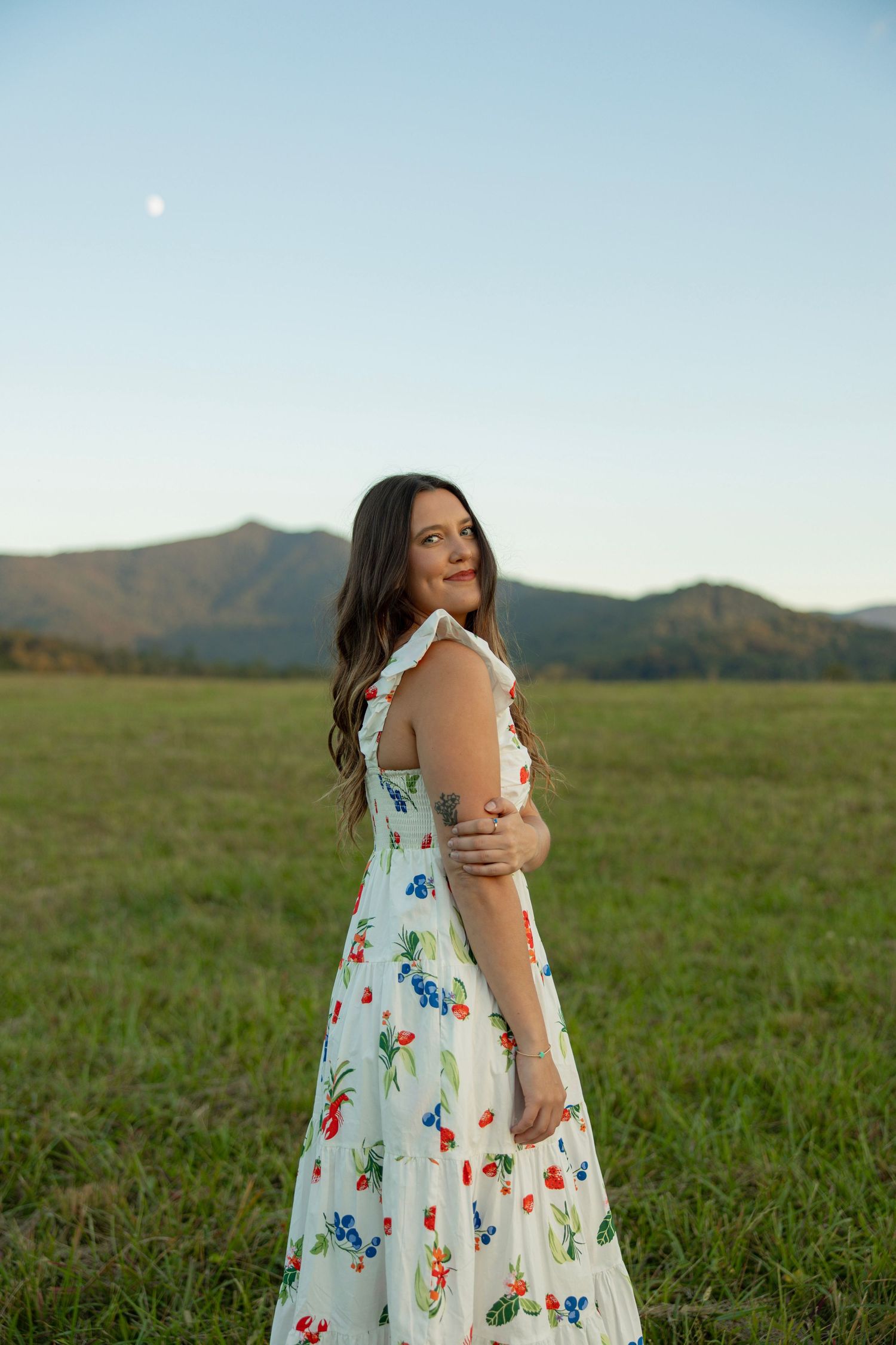 A woman in a floral print dress stands in a green field with mountains visible in the background.