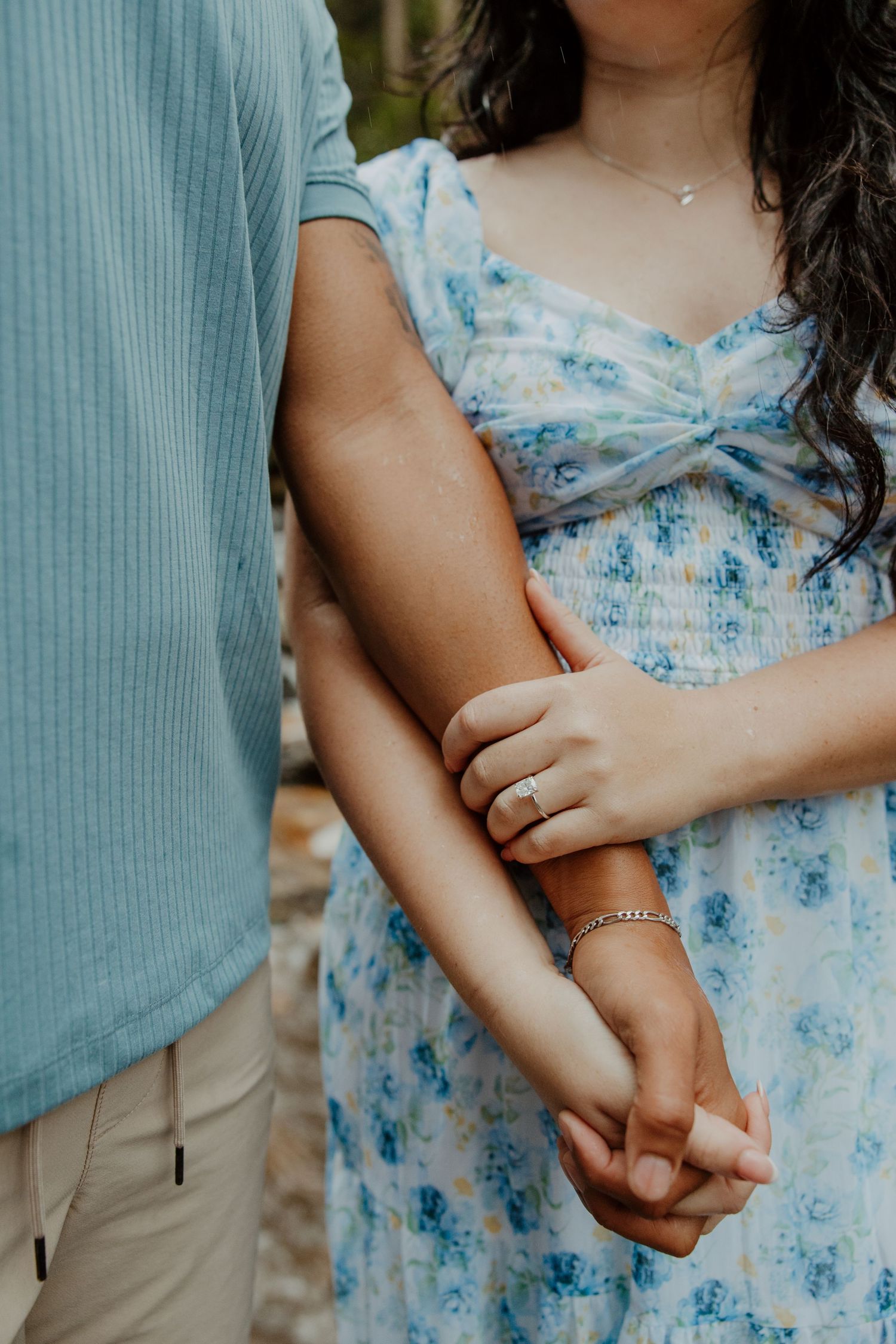 A romantic close-up of intertwined hands, with a blue floral dress and engagement ring visible.