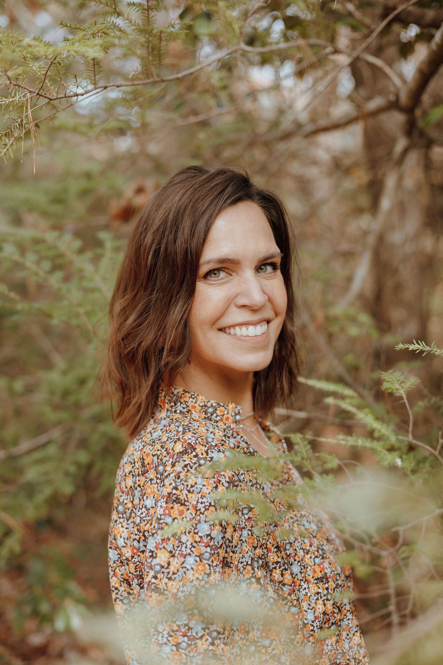 Portrait of a woman in a floral dress surrounded by natural foliage in warm lighting.