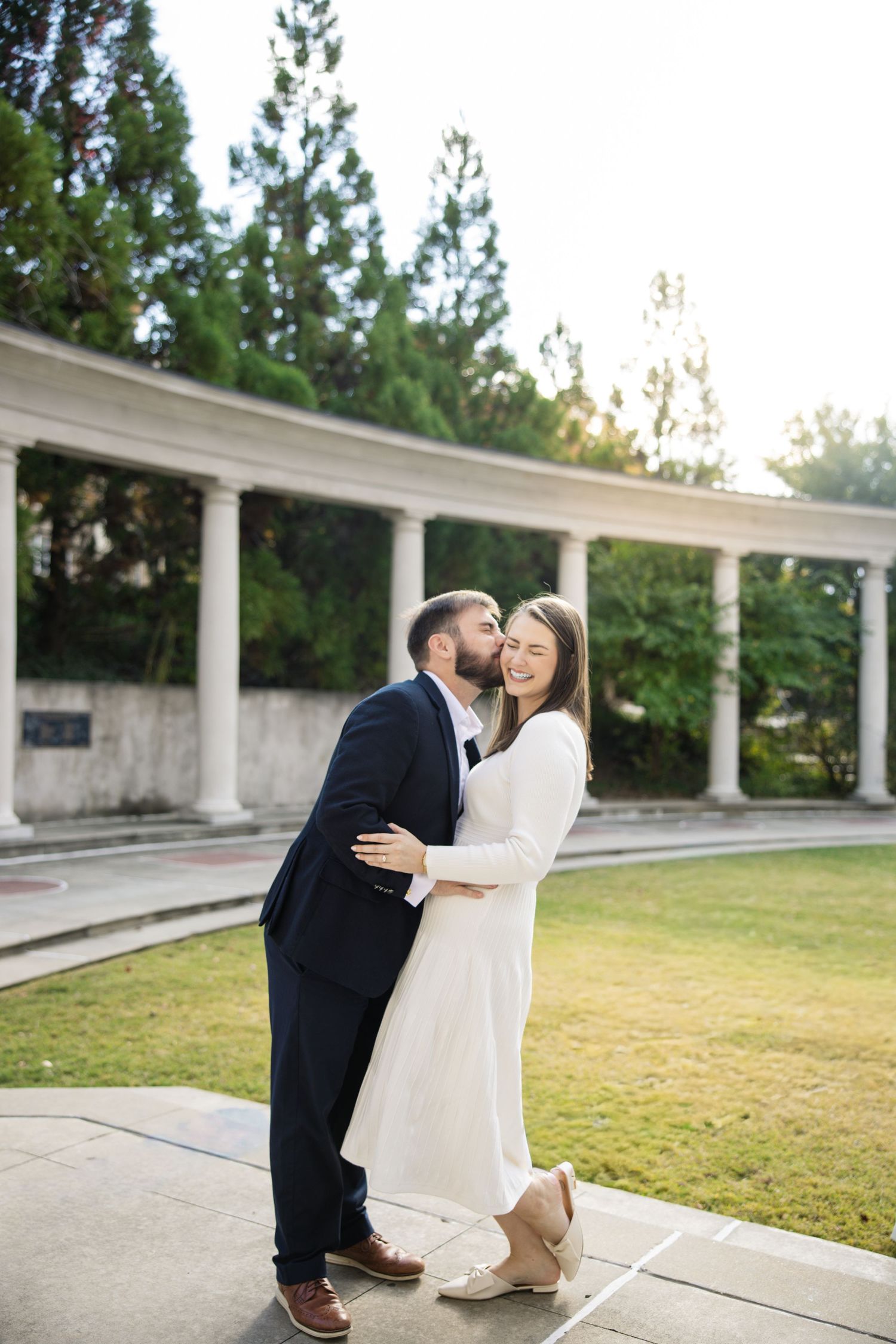 A romantic couple shares tender moments in a white columned outdoor setting during a photo session.