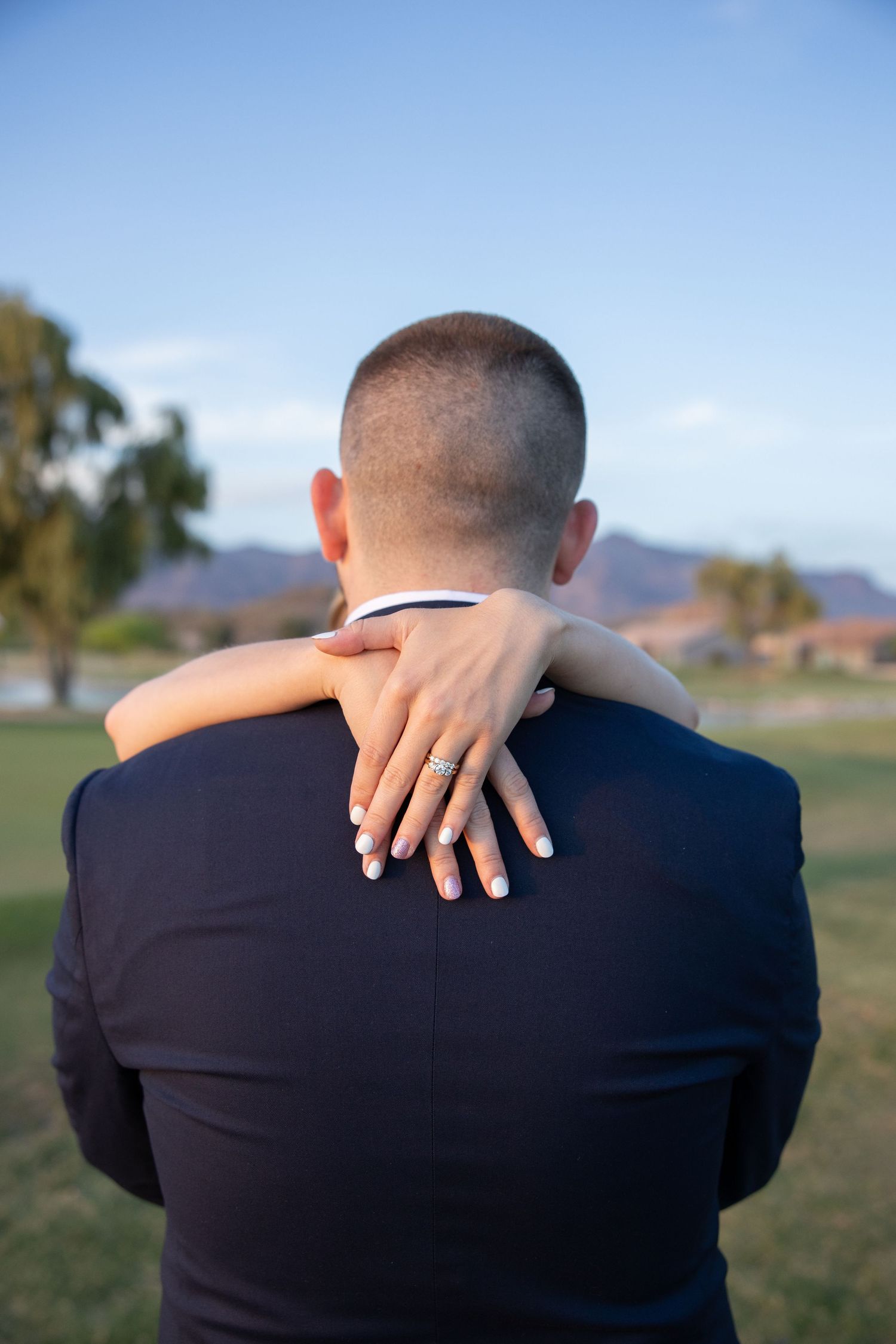 Close-up of bride's hand with diamond engagement ring resting on groom's back in navy suit outdoors.