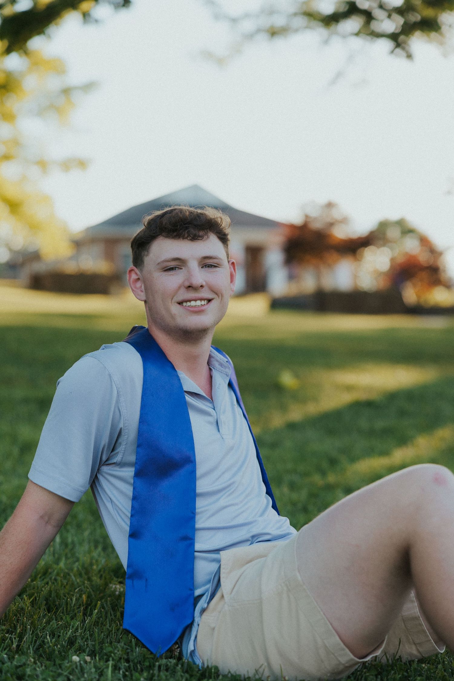 Graduate in blue stole sits on grass during sunset near campus building.