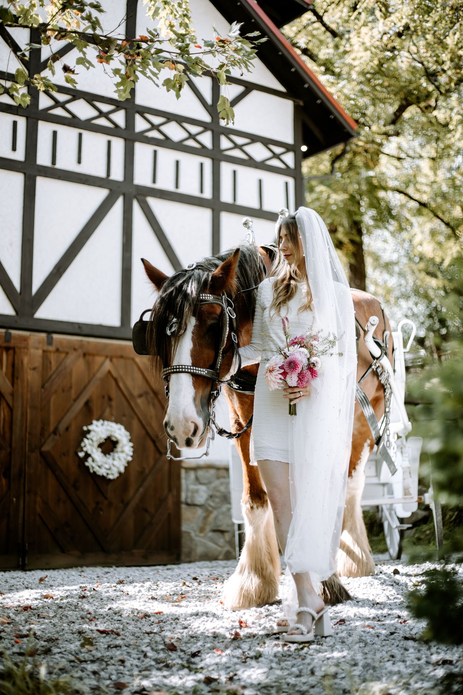 A white wedding dress and pink bouquet stand next to a brown and white horse against a Tudor-style barn with wreath decor.