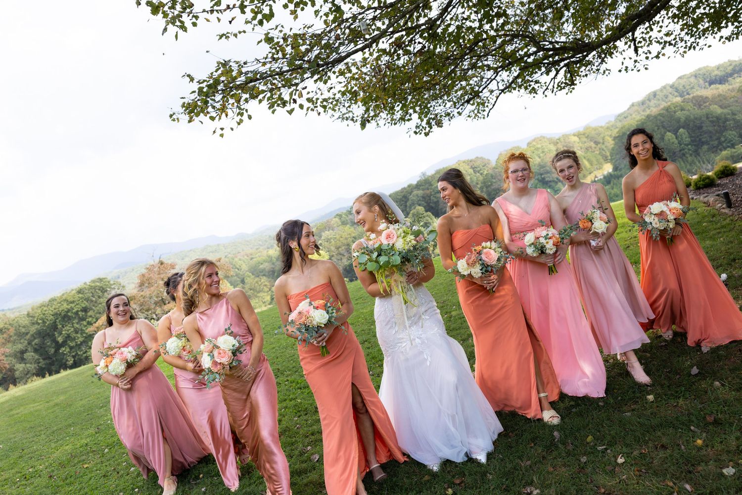 Wedding party in coral and pink dresses poses together on a grassy hillside for group photos at sunset.