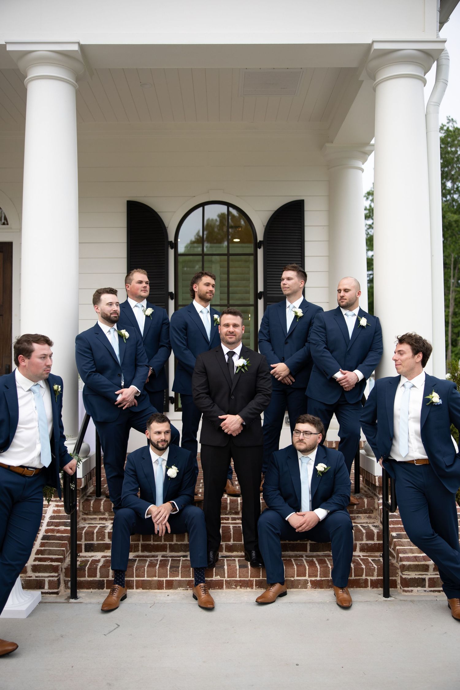 Groom in dark suit stands centered among groomsmen in navy suits on white columned venue steps for wedding party photo.