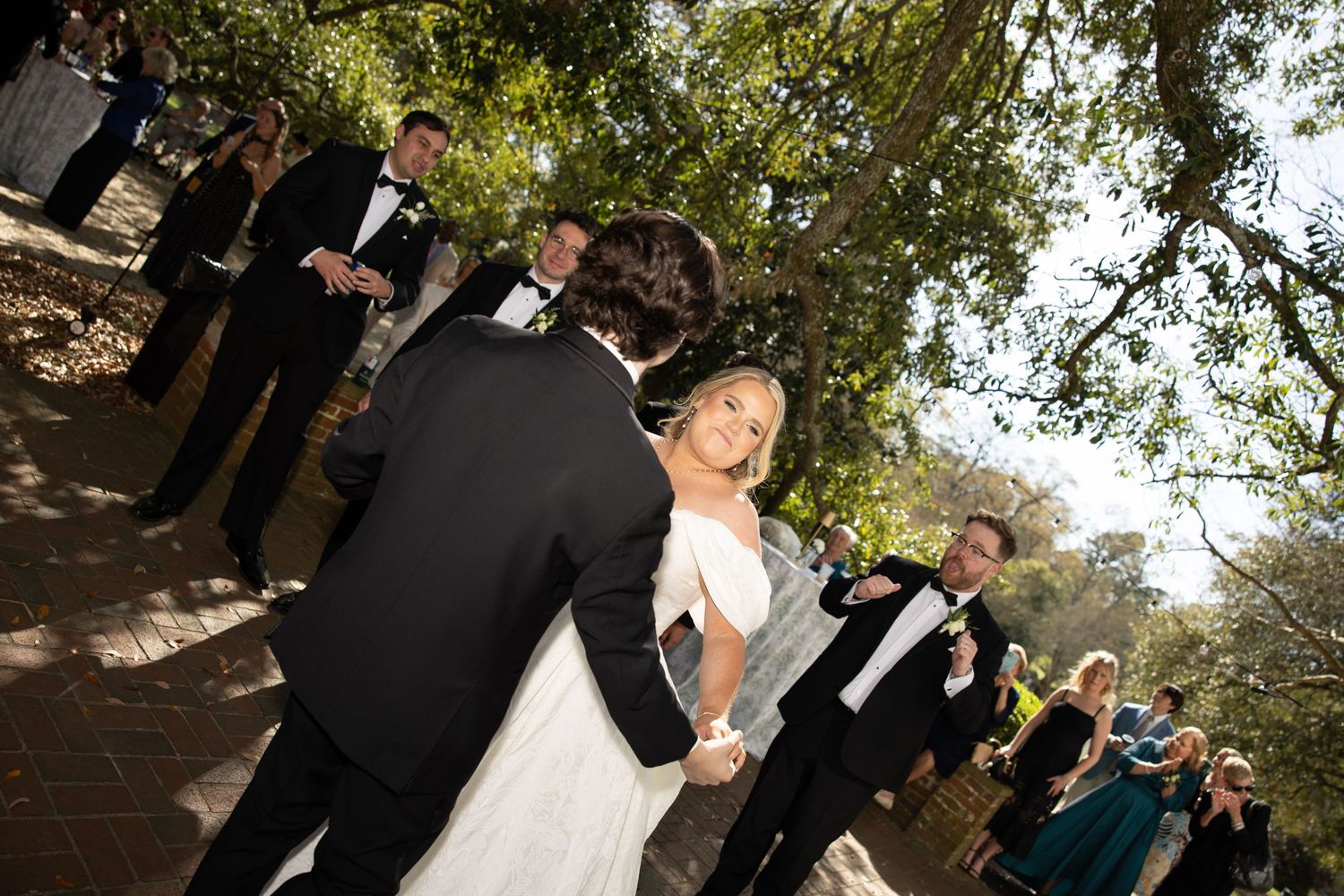 Bride and groom share first dance outdoors at elegant wedding ceremony surrounded by guests under shady trees.