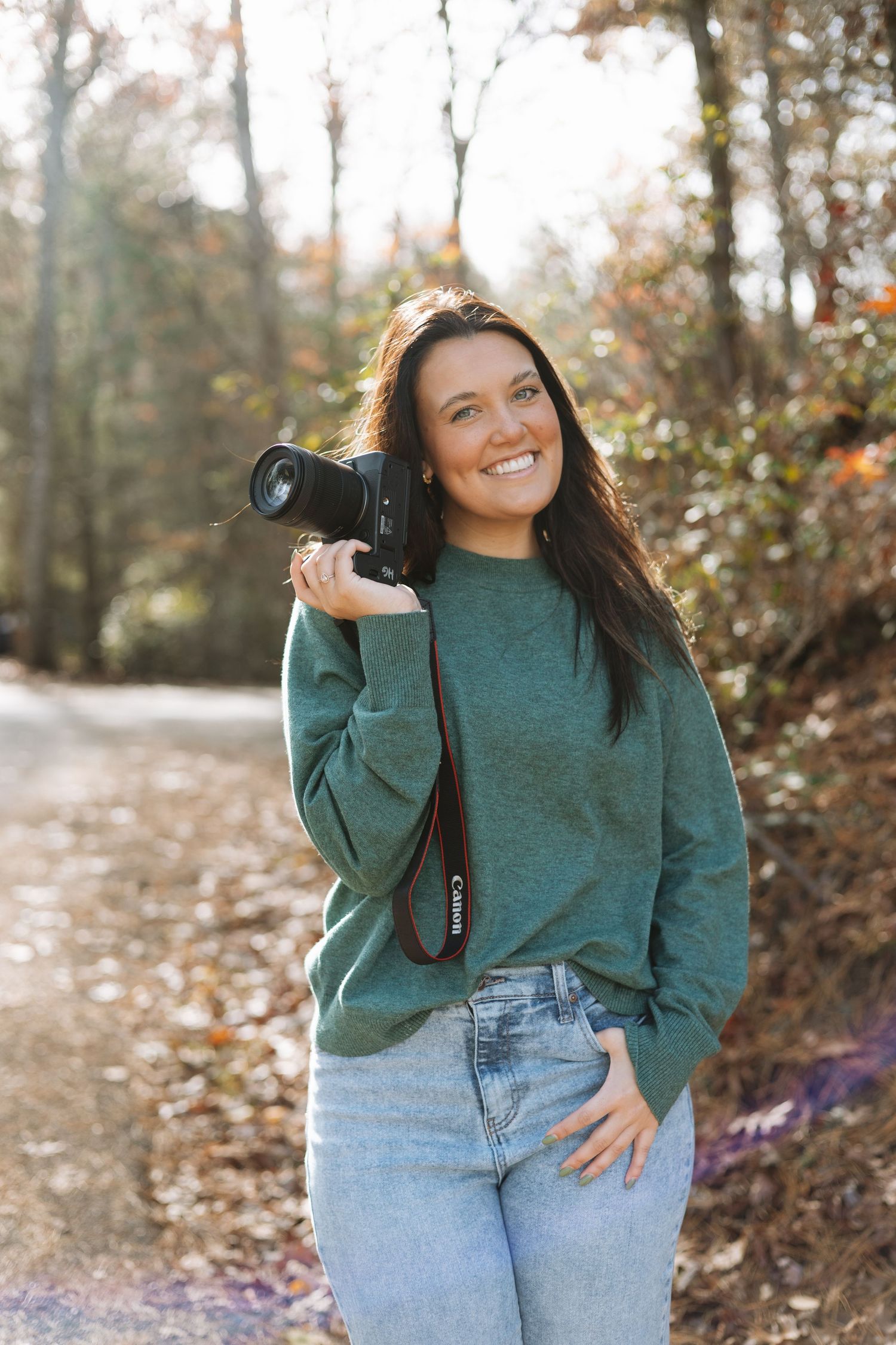 Photographer in green sweater and jeans holding camera on autumn trail.