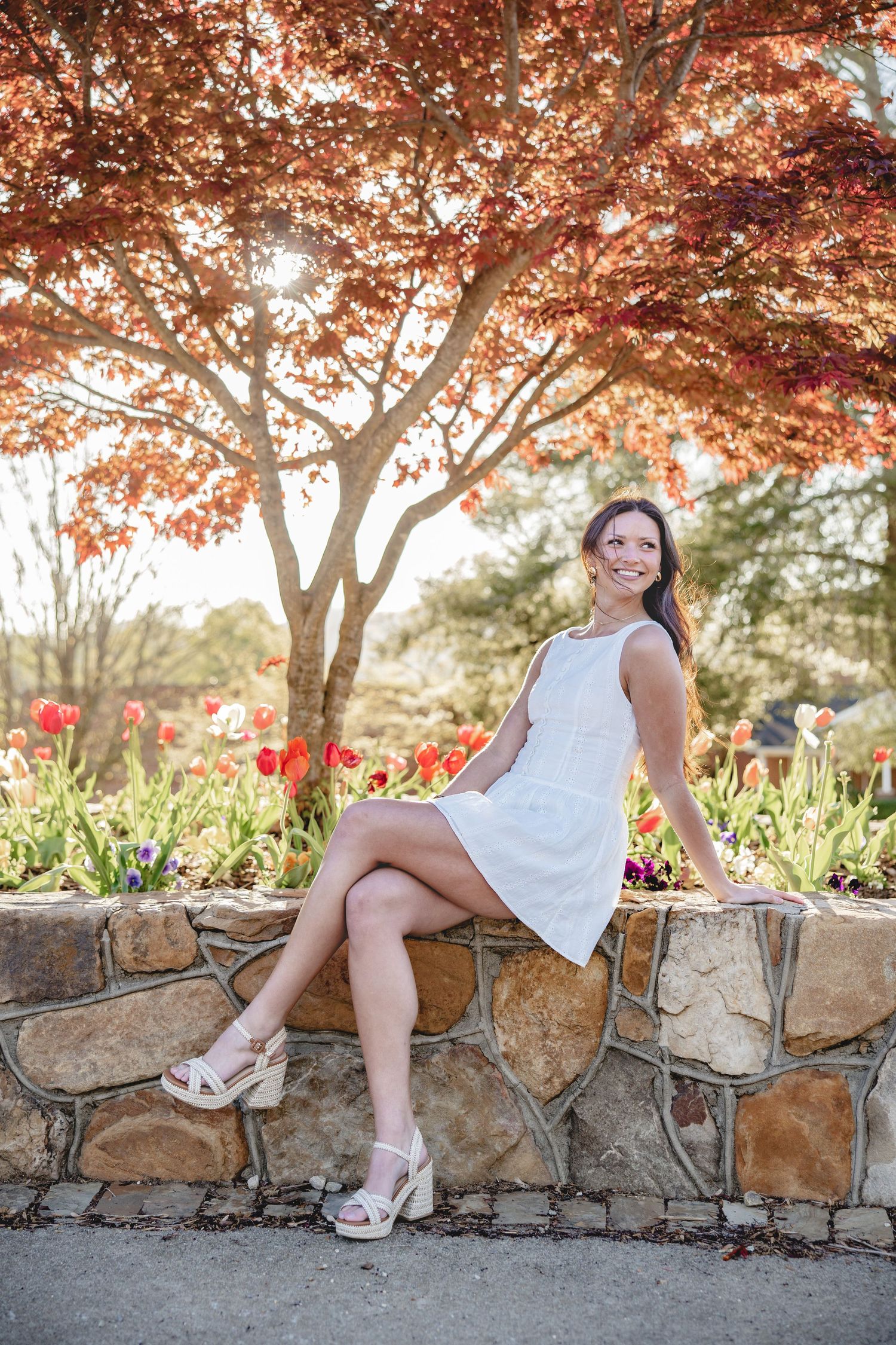 Graduate posing by stone wall under blooming autumn trees with red flowers.