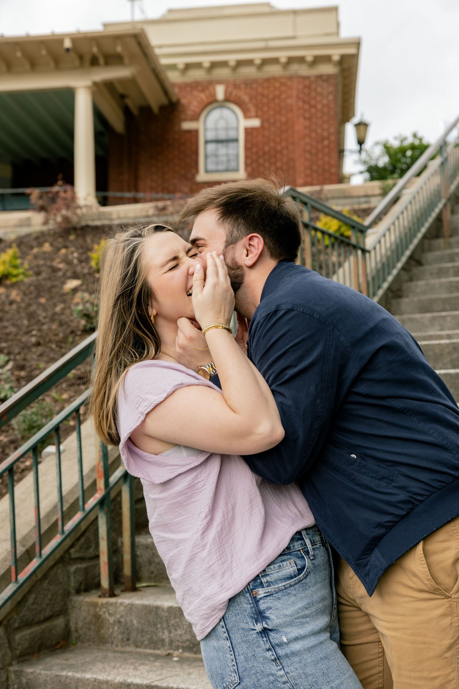 A romantic couple shares an intimate moment on stone steps near a brick building.