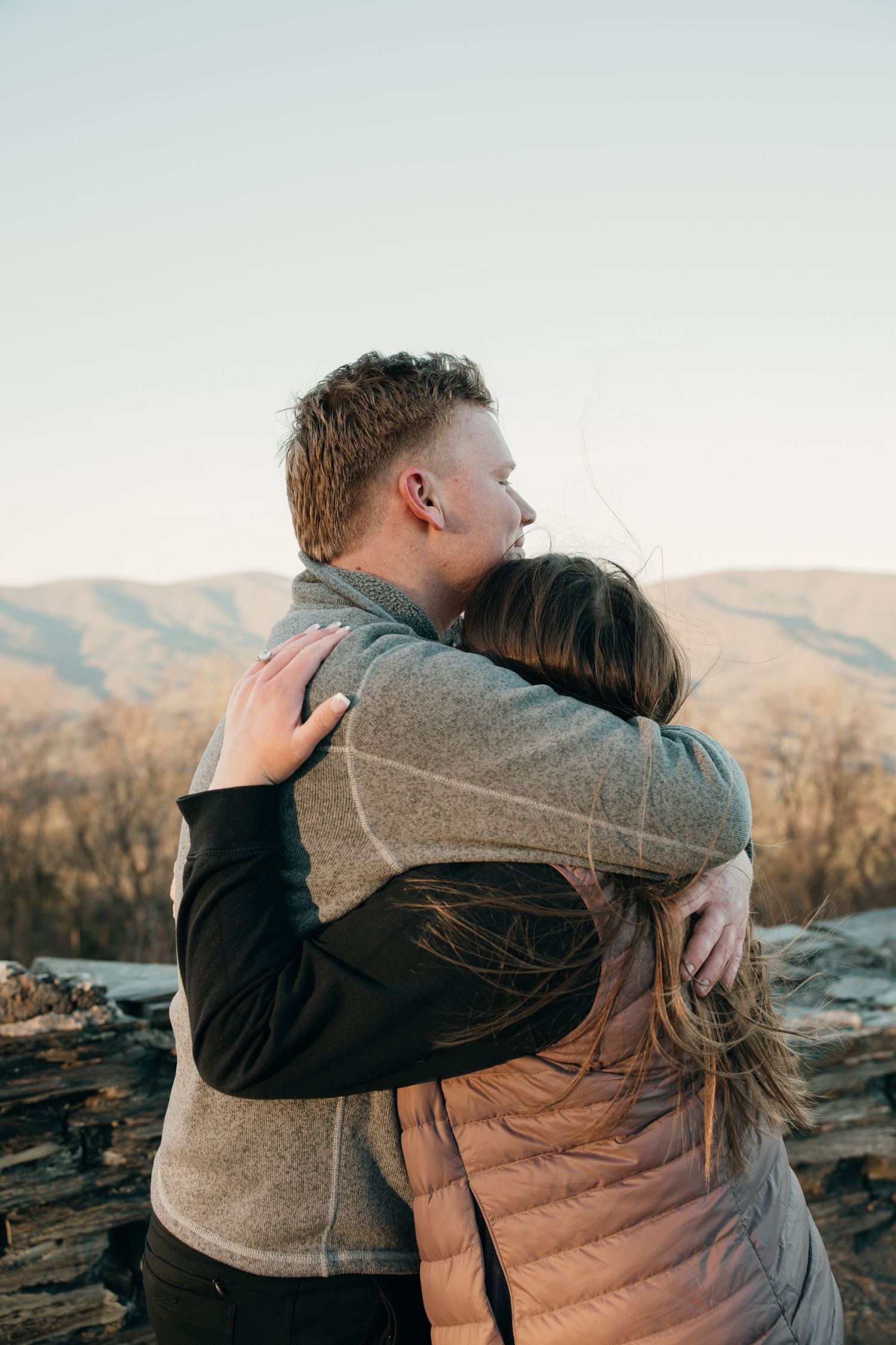 A romantic couple embraces while sitting on a mountain overlook during sunset.
