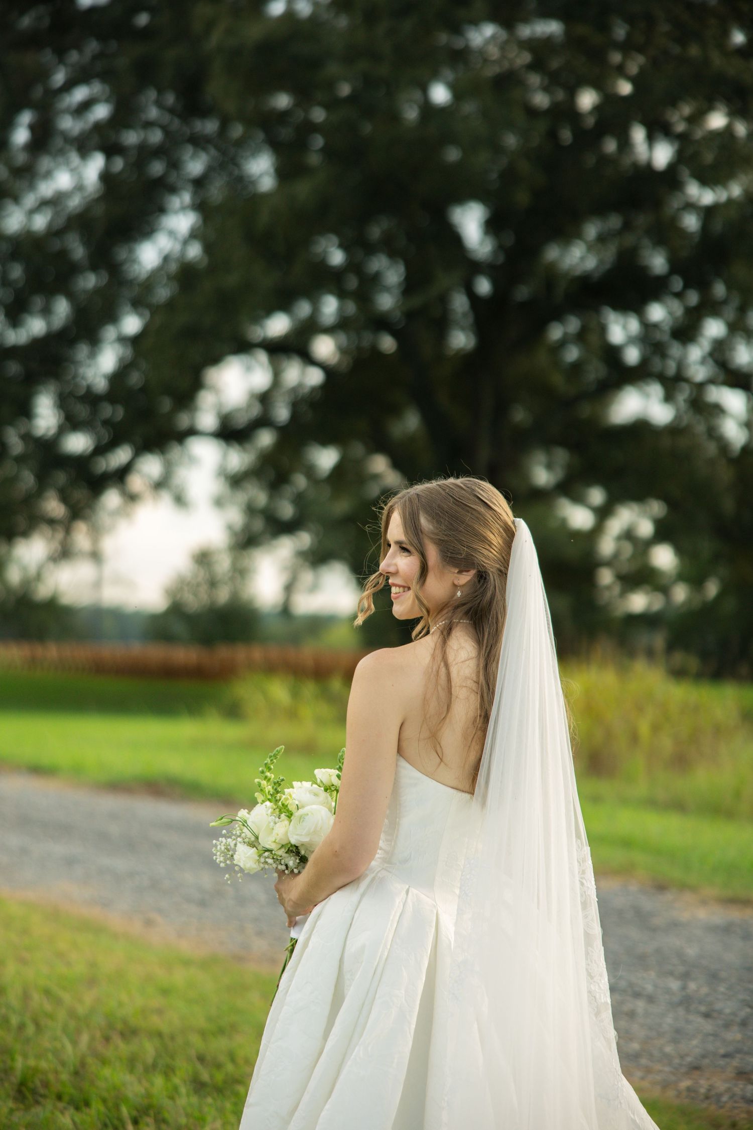 A bride in a flowing white wedding dress and veil stands on a country path with trees and green fields in the background.