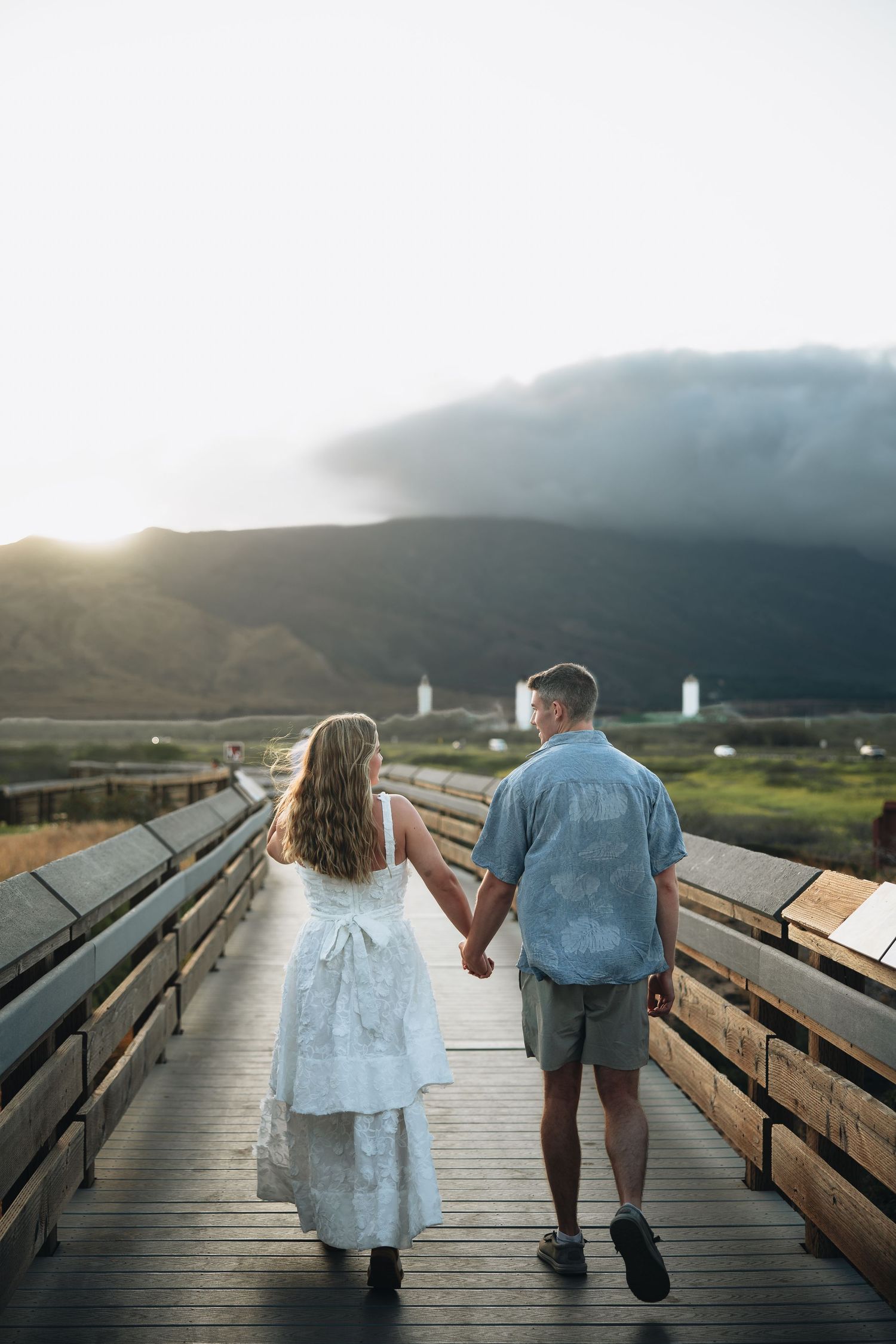Couple walking hand in hand on a wooden boardwalk at sunset with mountains and dramatic skies in the background.