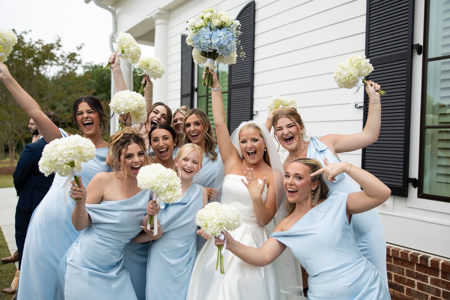 Bride and bridesmaids in light blue dresses celebrate joyfully holding white bouquets outside a white building.