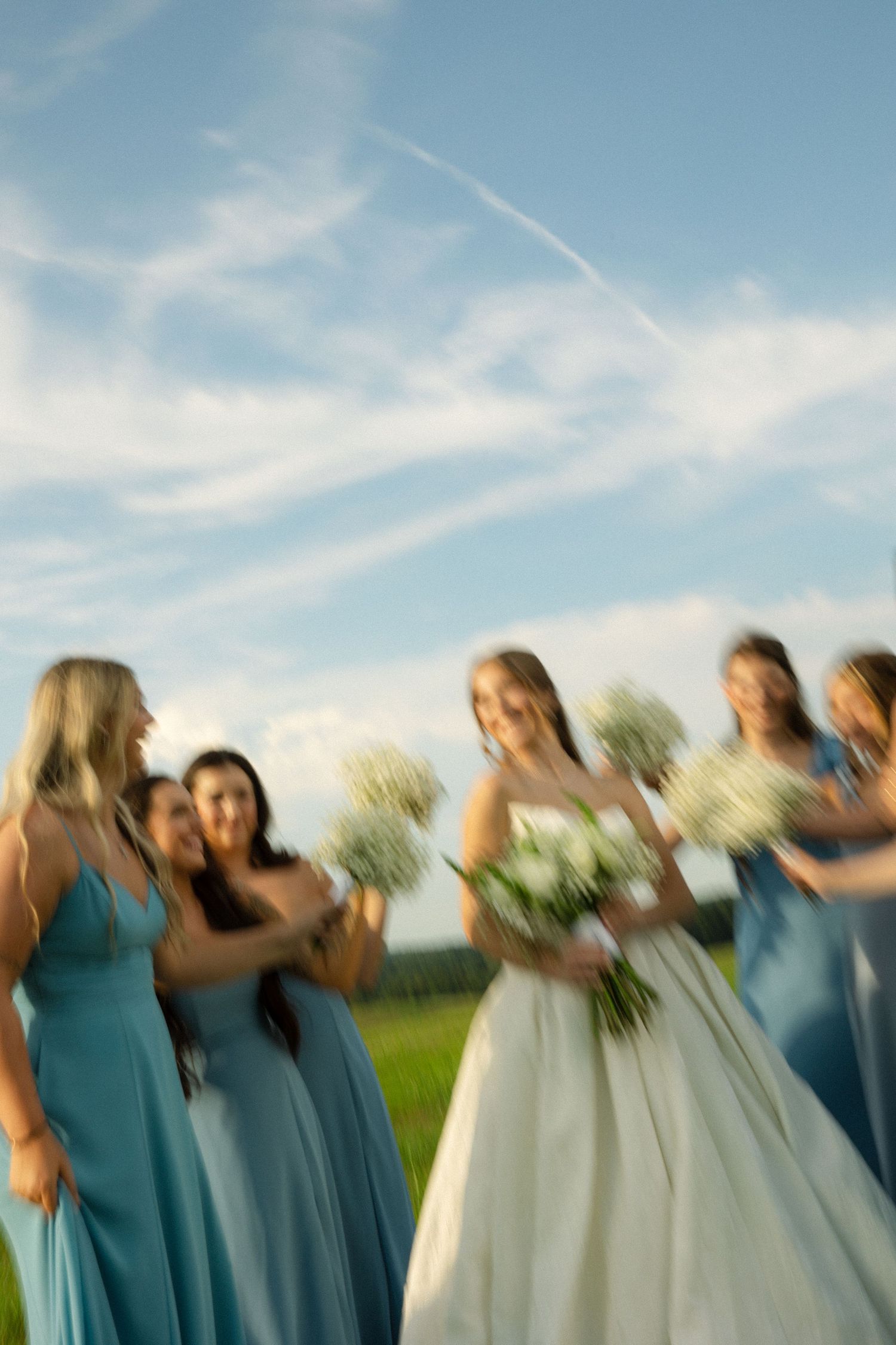 A group of bridesmaids in blue dresses dance and twirl together in a joyful motion blur on a sunny outdoor day.