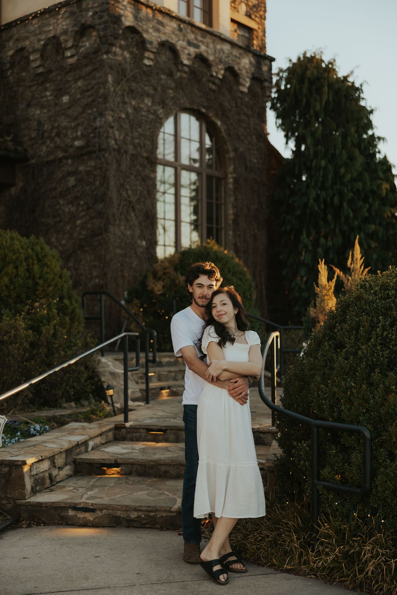 A romantic couple embraces on stone steps outside a historic stone building with arched windows at sunset.