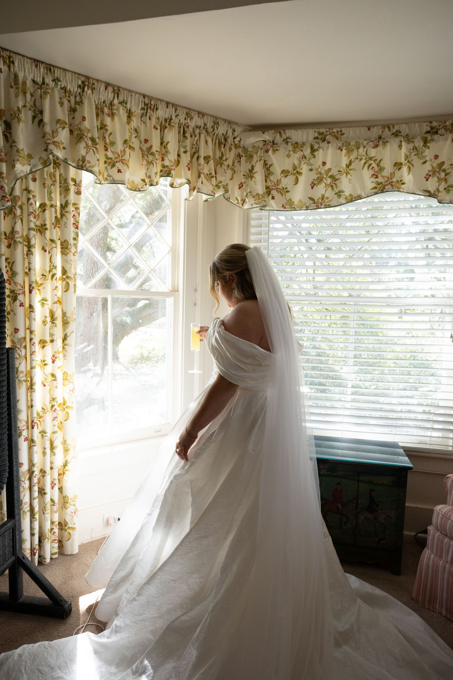 A bride in a flowing white wedding gown twirls and dances in front of a window with floral curtains.