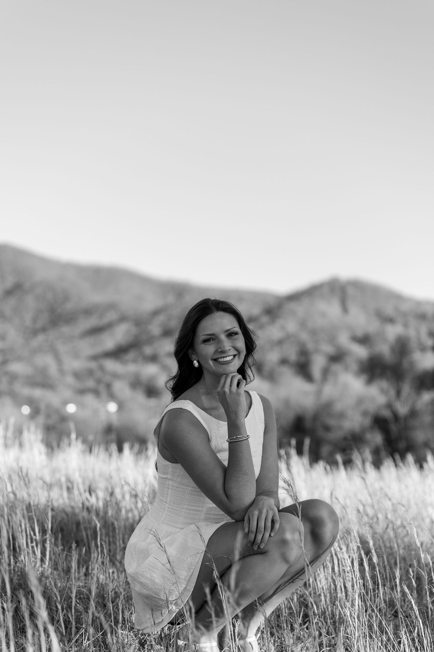 Person in white dress poses in tall grass with mountain backdrop in black and white.