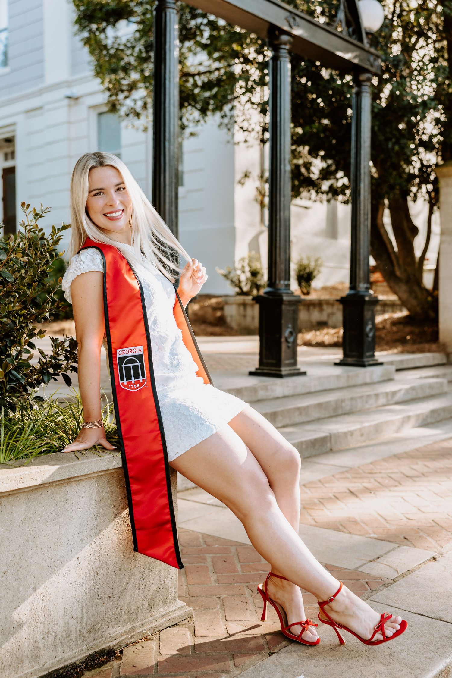 Graduate in white dress and red graduation stole poses on campus steps.