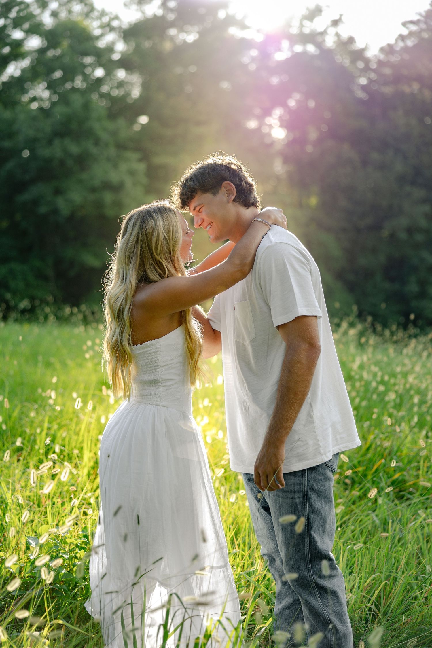 A couple in white clothing embraces romantically in a sunlit meadow with tall grass and wildflowers.