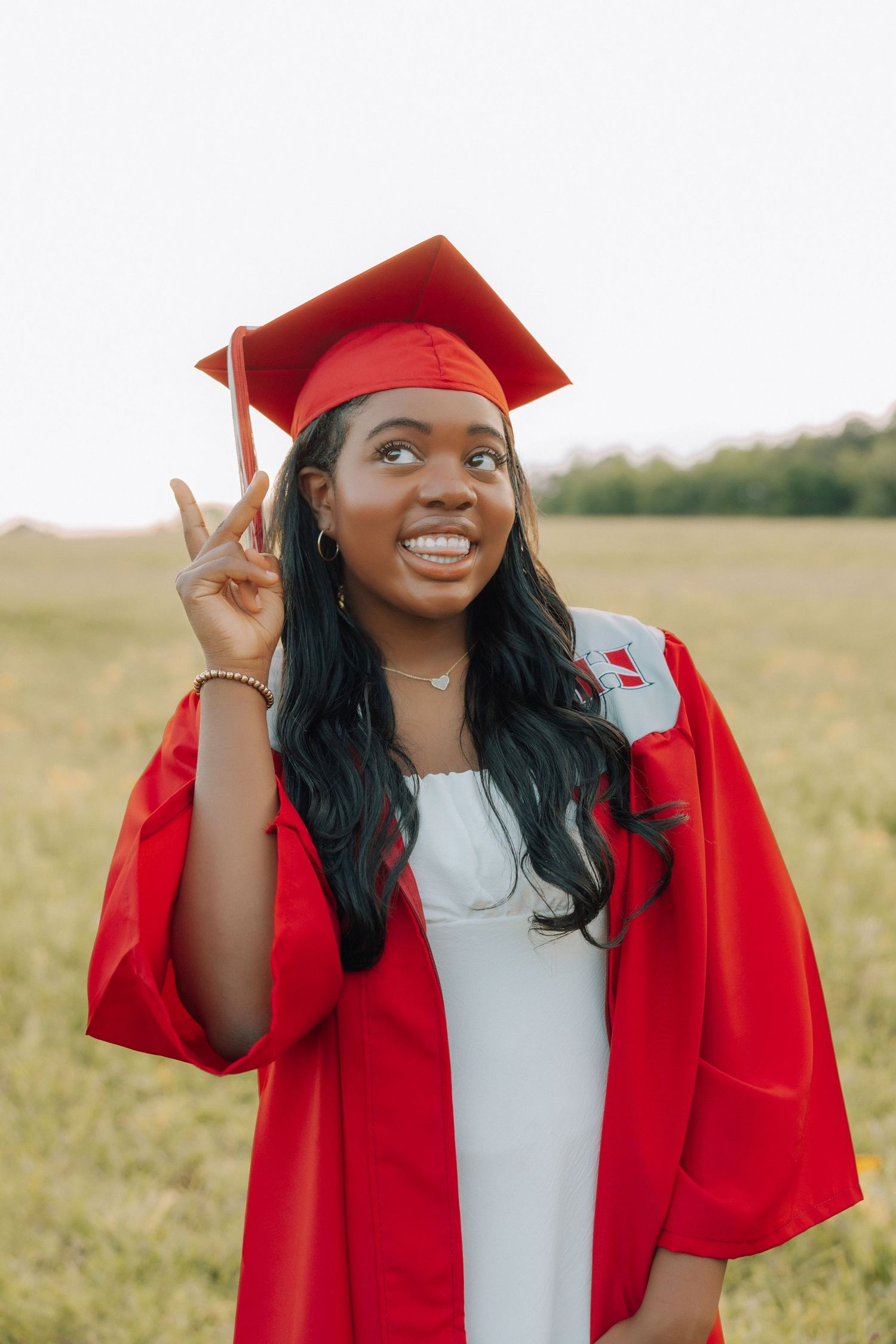 Graduate in red cap and gown makes peace sign while smiling outdoors.