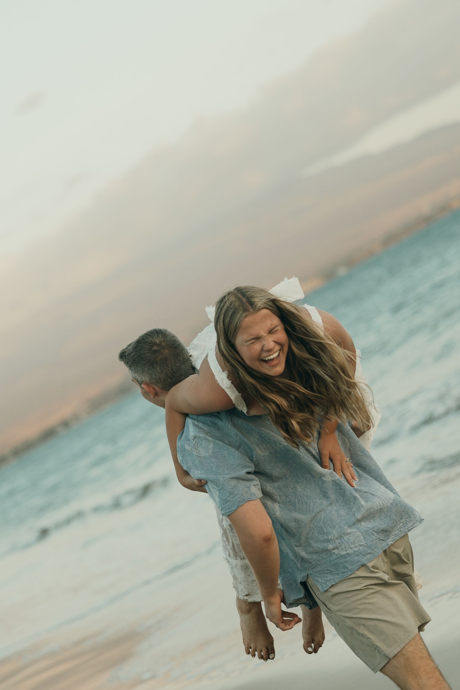 A person in denim clothing runs and jumps joyfully along a beach at sunset with waves in the background.