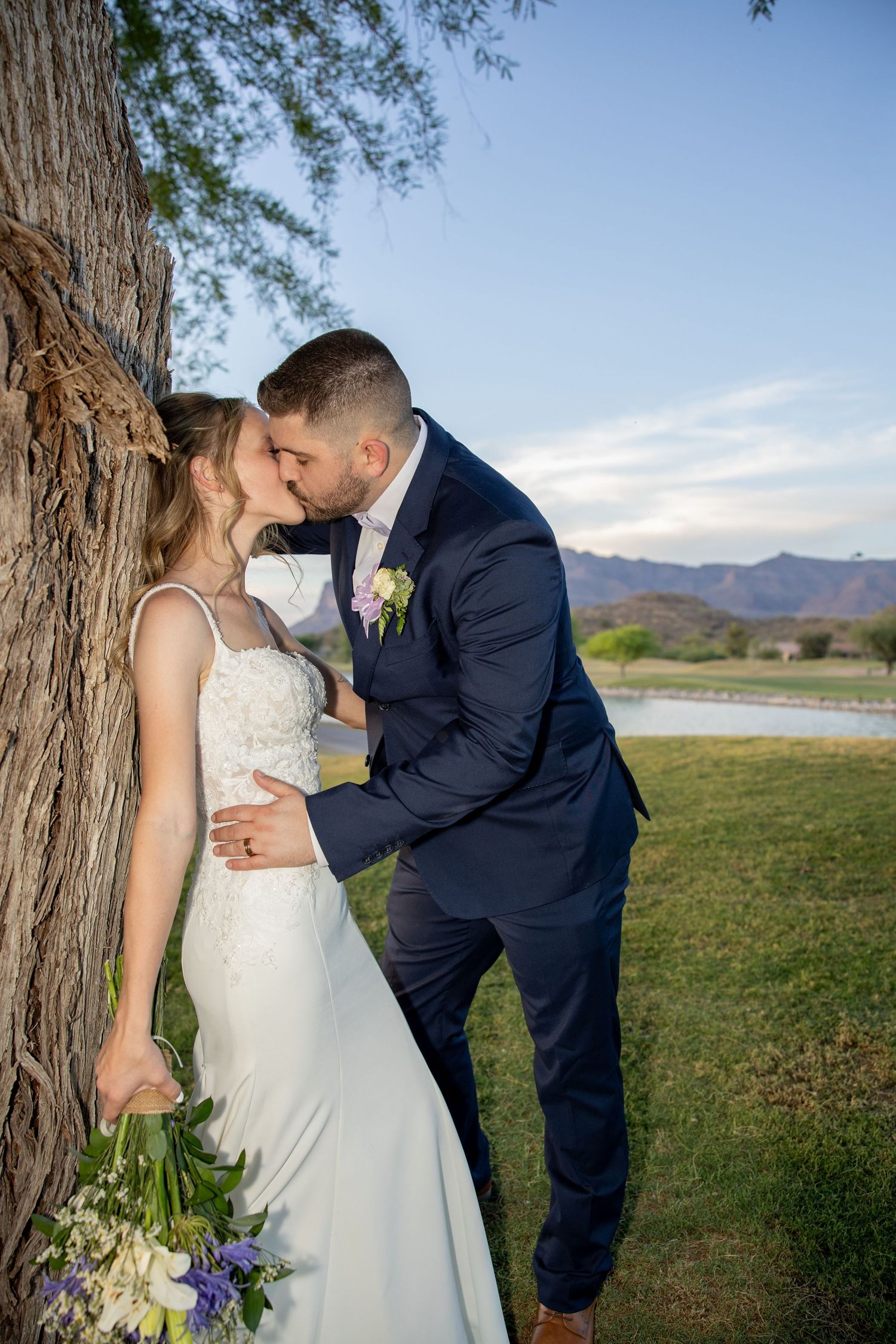 Bride and groom sharing a kiss beside a large tree with mountains and blue sky in the background.