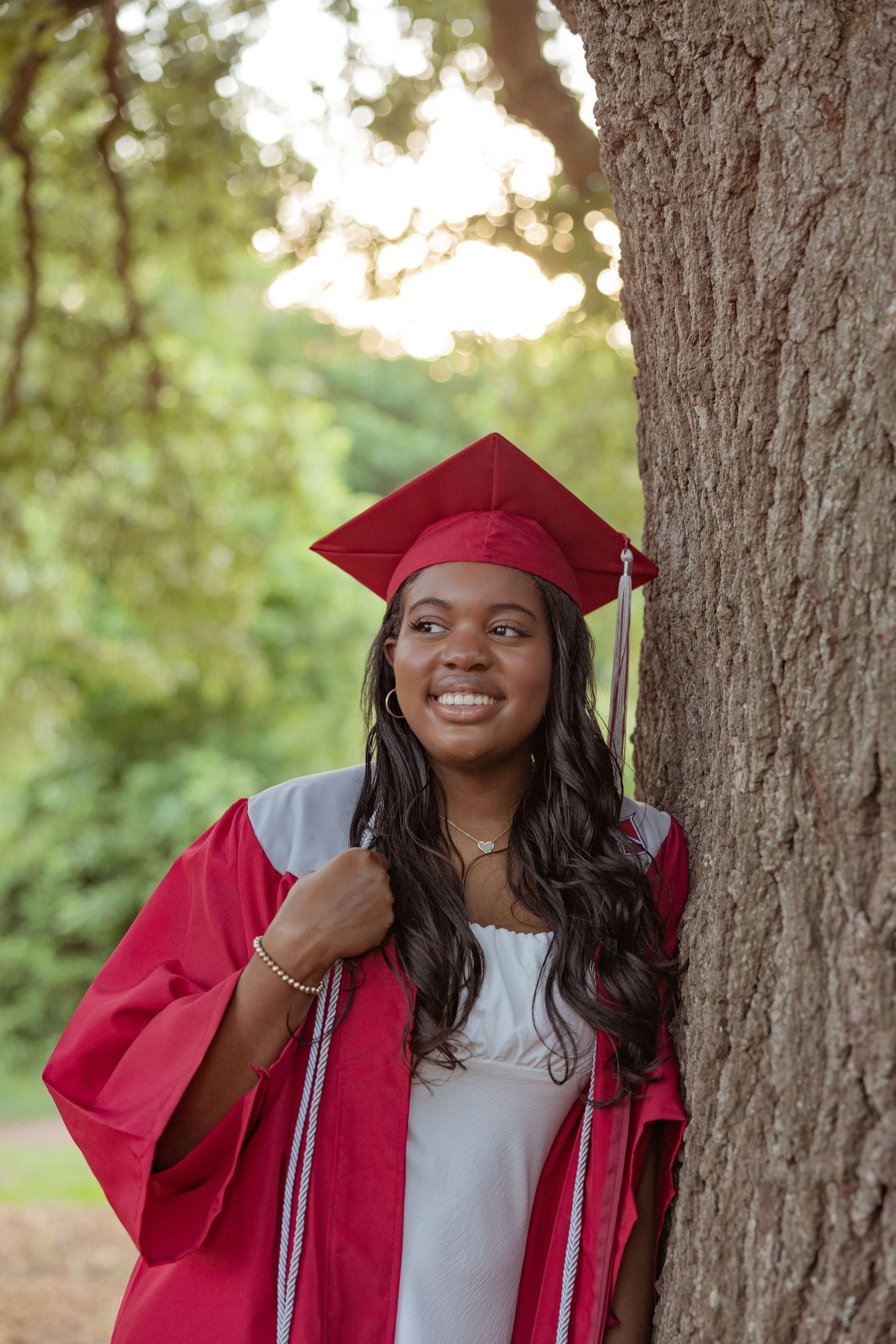 A graduate in red regalia smiles while standing next to a large tree trunk.