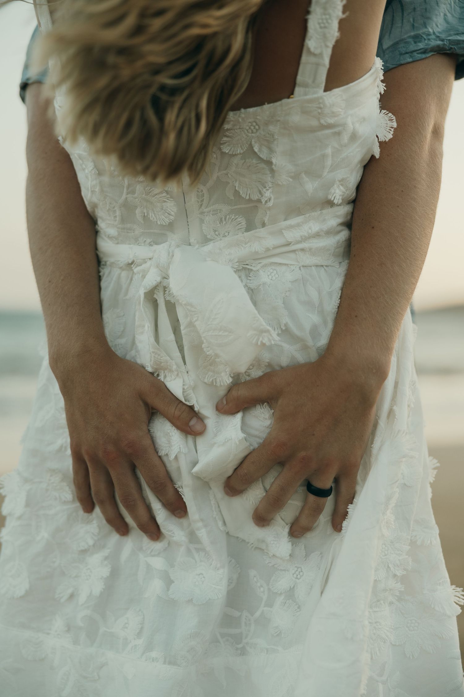 Close-up view of hands resting on a white lace wedding dress with delicate embroidery and a black ring detail.