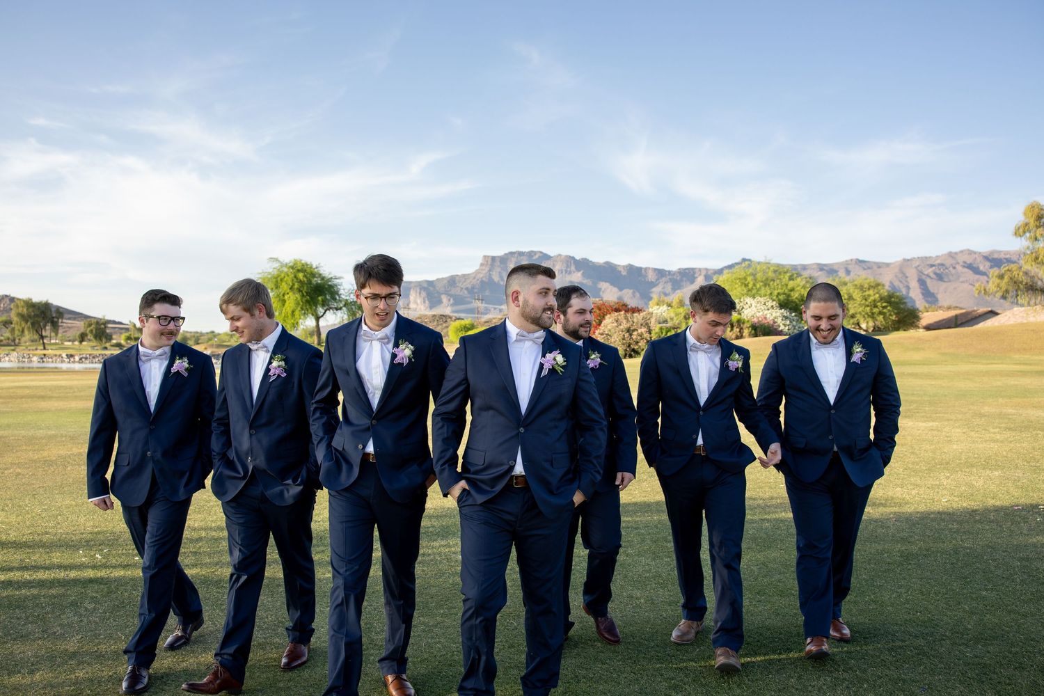 Groom and groomsmen in navy suits walking outdoors on green grass with mountain backdrop at a sunny wedding.