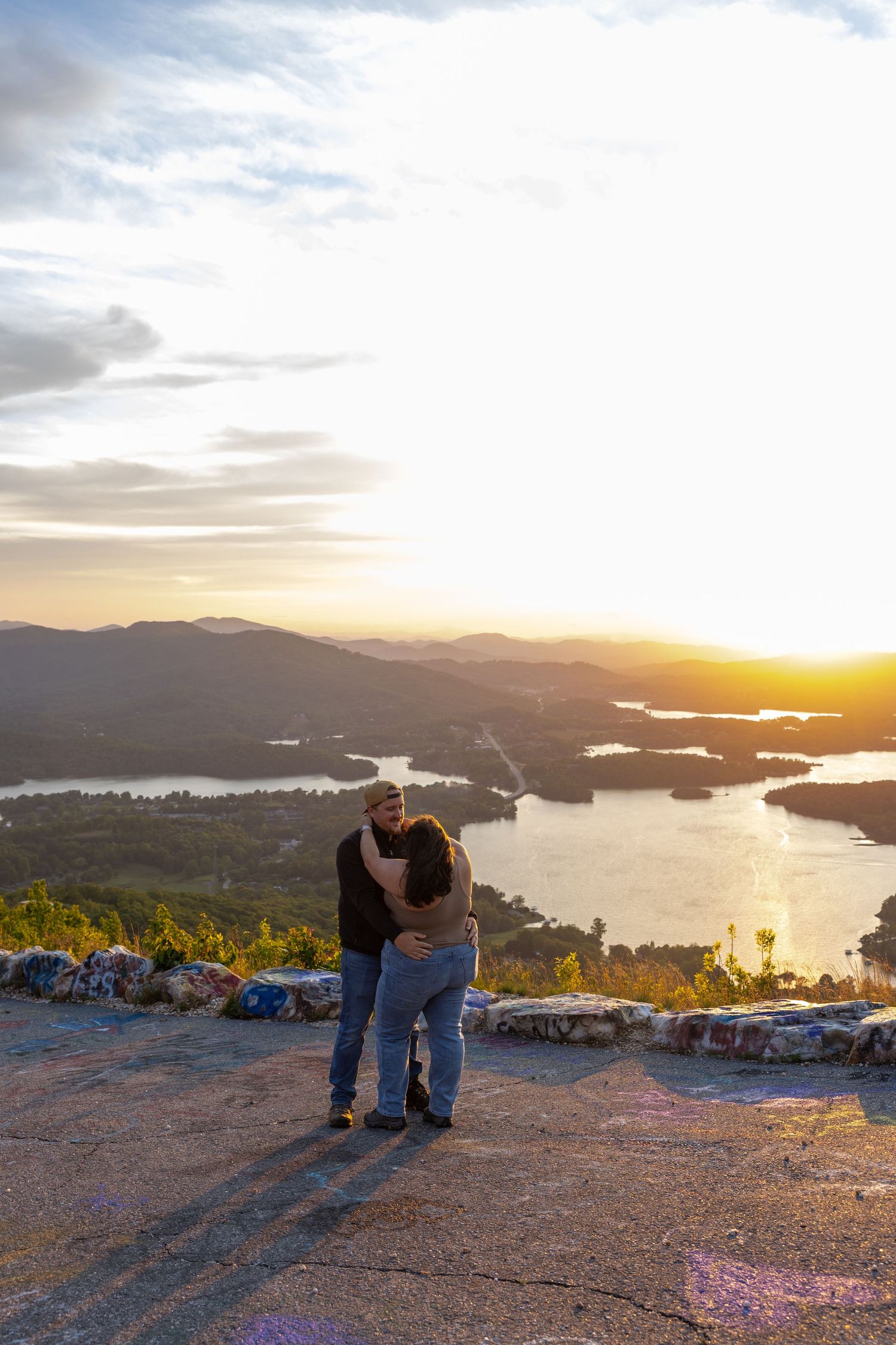 Couple embracing at sunset on a mountain overlook with panoramic views of a lake and rolling hills below.