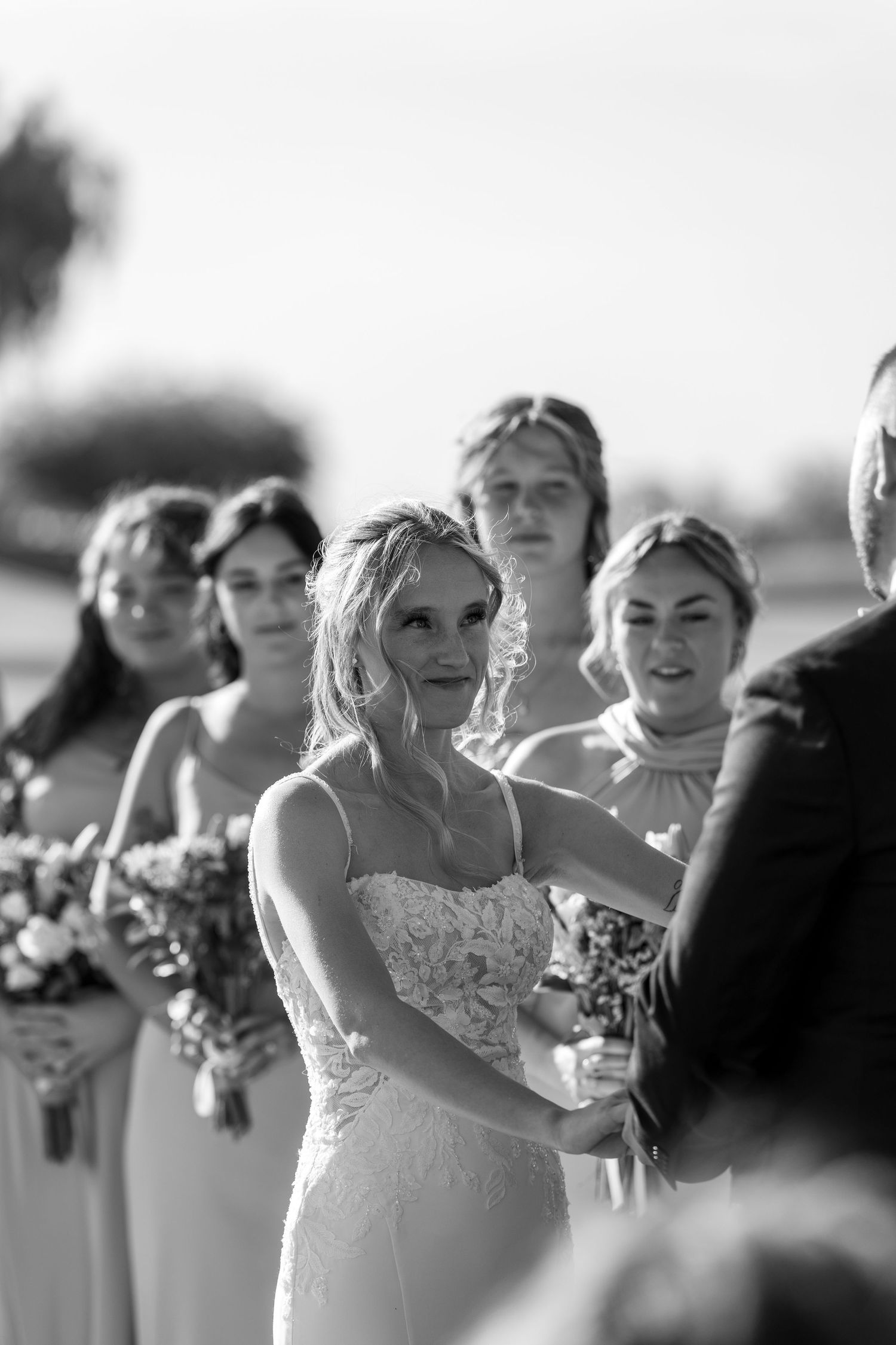 Black and white photo of a smiling bride in lace gown holding hands at outdoor wedding ceremony with bridesmaids behind her.