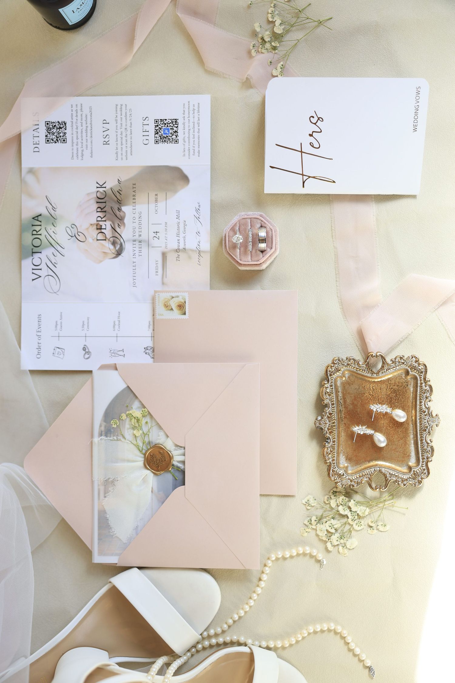 Elegant wedding flat lay with blush pink envelopes, invitation suite, pearl necklace, gold tray with earrings, and bridal accessories.