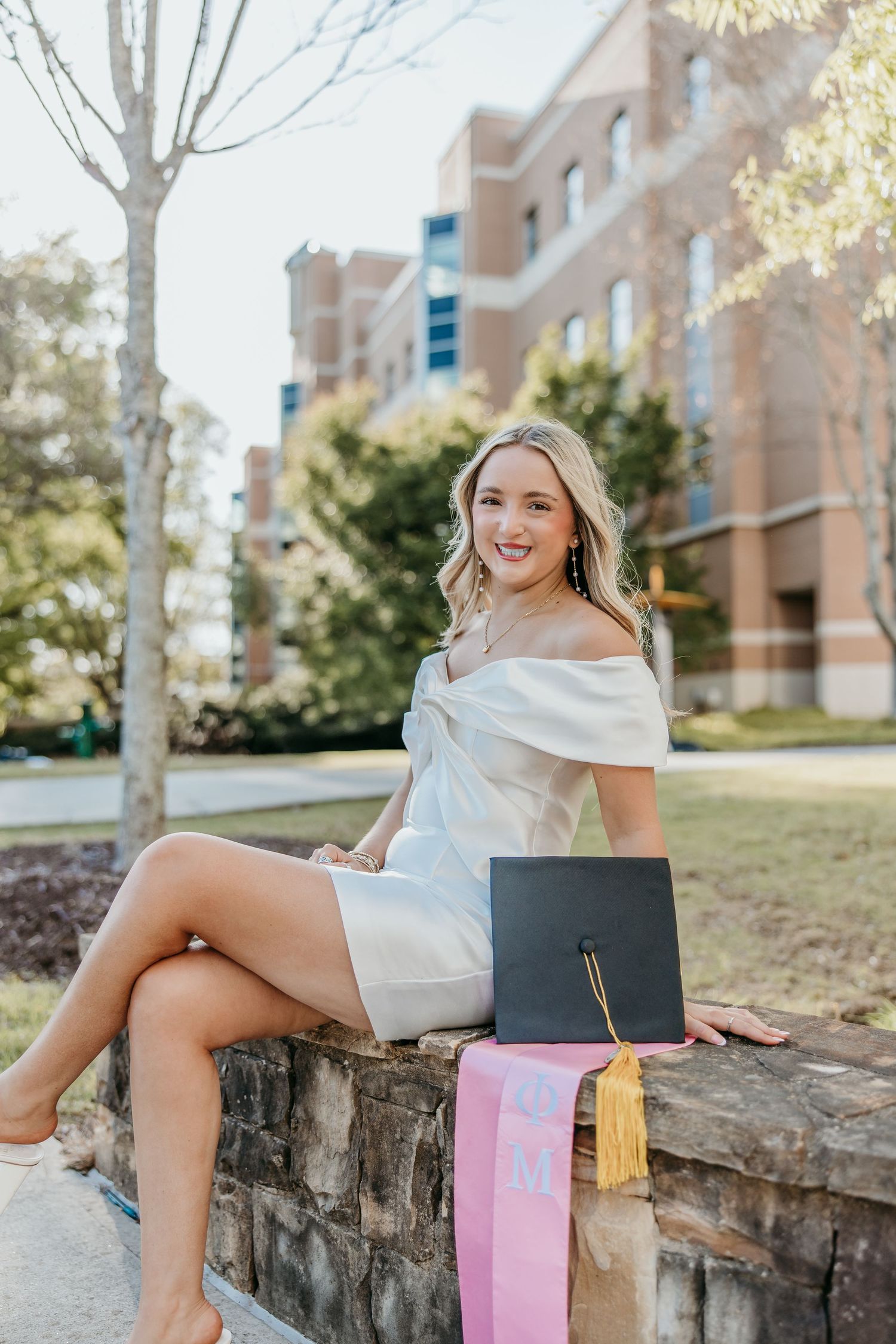 Graduate in white dress sits on stone wall with graduation cap and pink stole.