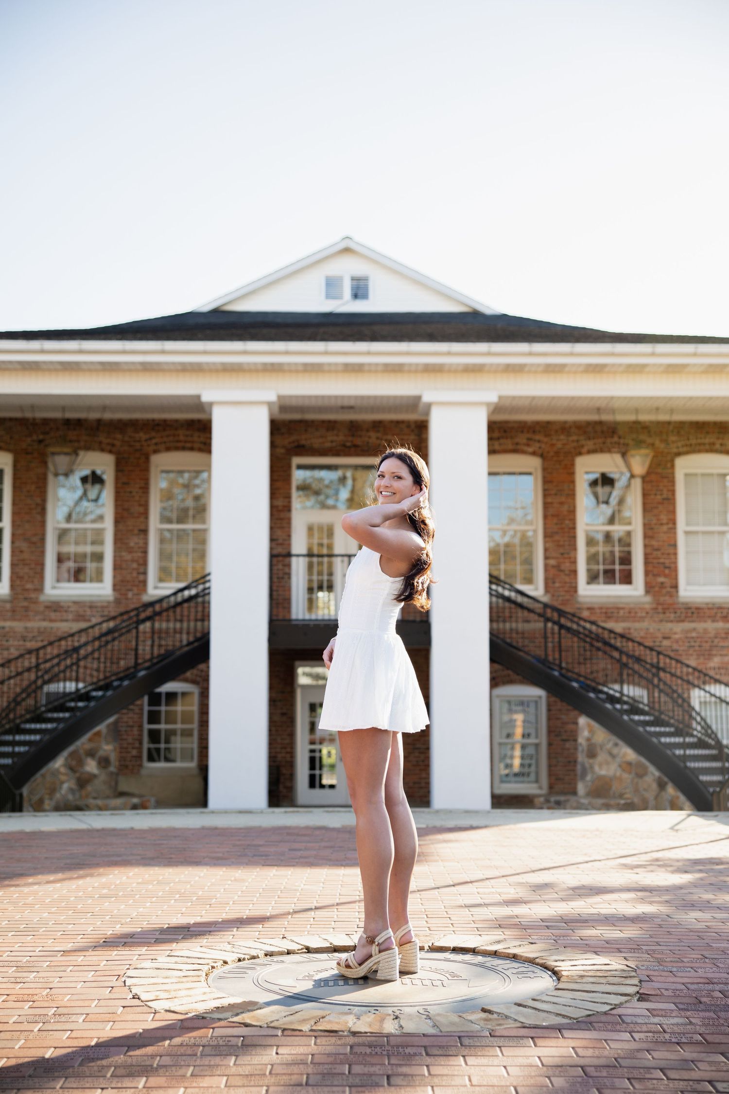 Graduate in white dress posing on brick courtyard in front of columned building.