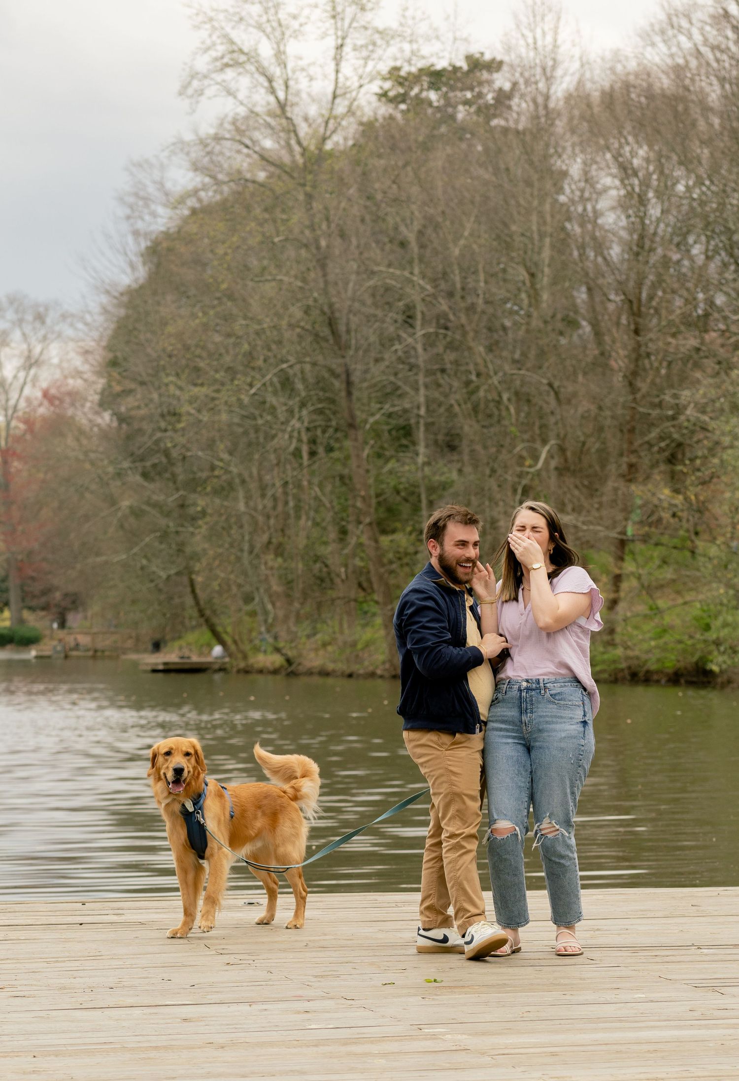 A happy couple celebrates on a lakeside dock with their golden retriever companion.