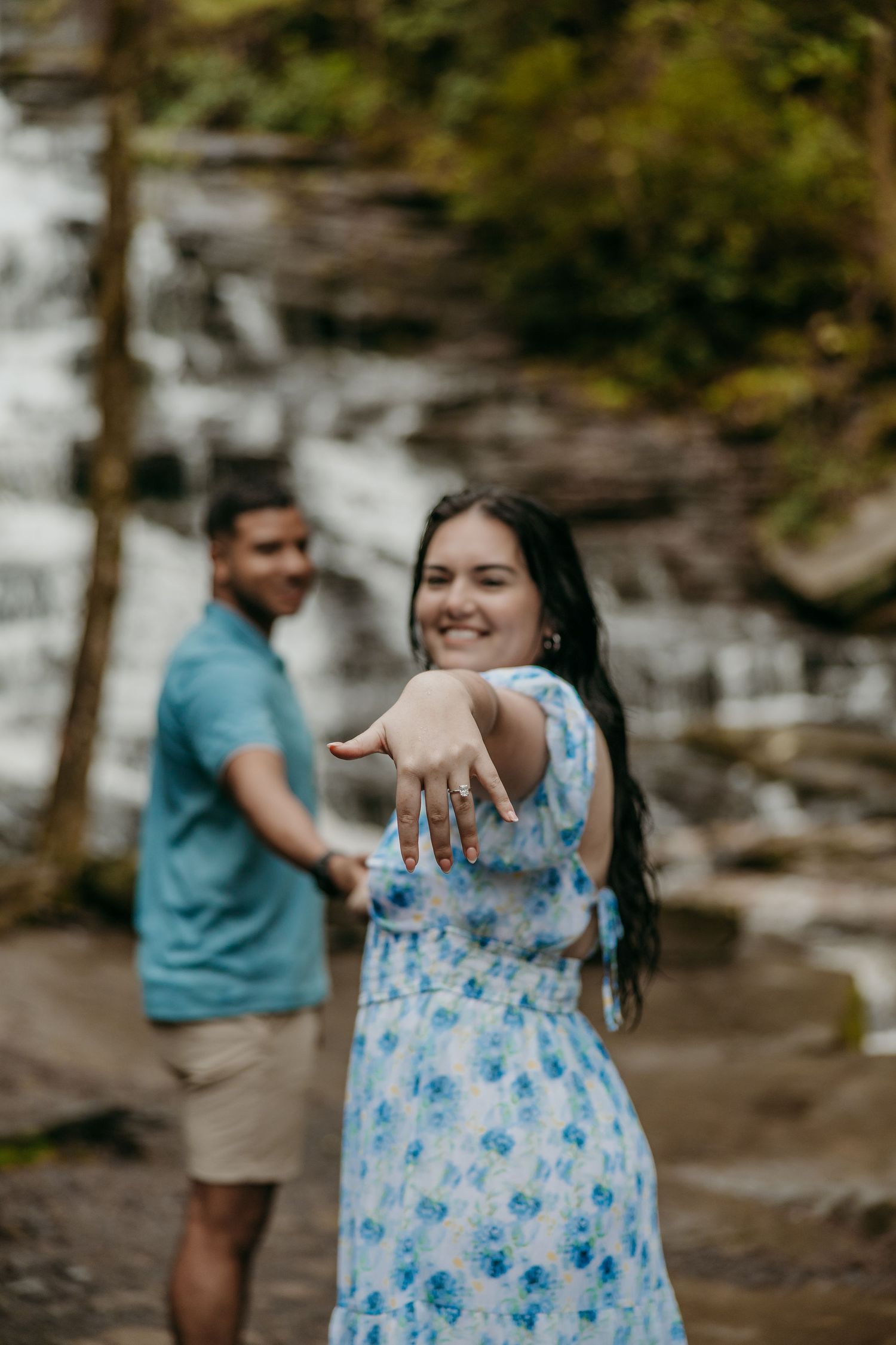 A couple stands by a waterfall while showing off an engagement ring in a romantic outdoor proposal photo.