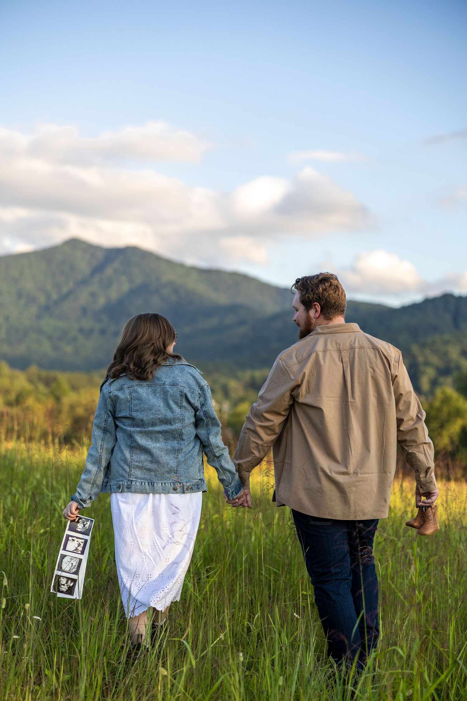 Couple walking hand in hand through a green meadow with mountains in the background holding a sign.