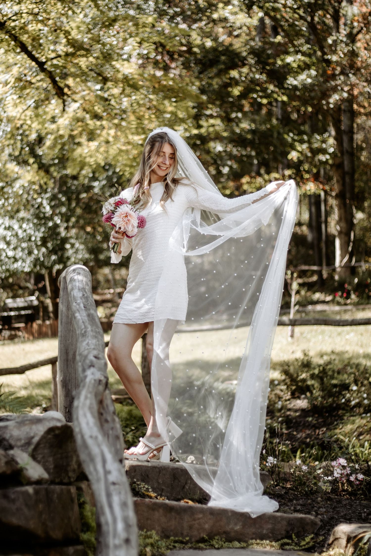Bride in short white dress poses with flowing veil and pink bouquet in garden setting.